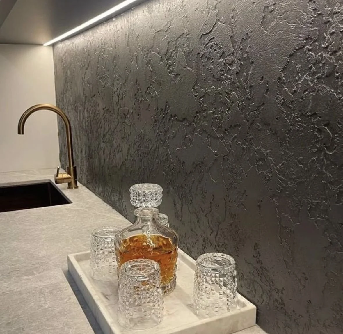 A kitchen countertop with a gold-colored faucet, a bottle of liquor with a glass stopper, and four textured glass tumblers on a white tray. The background features a dark textured wall with a strip of LED lighting above.