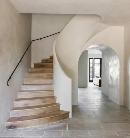 Interior view of a minimalist home with a curved staircase featuring wooden steps and a black handrail, adjacent to an arched doorway leading to a bright room with large windows.