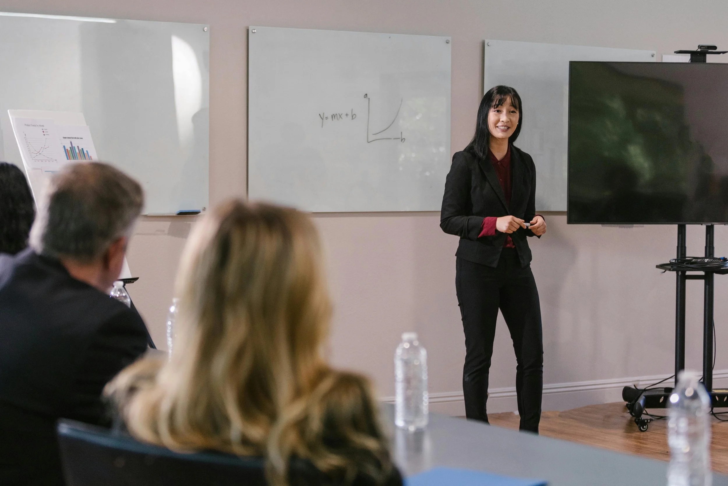 A woman standing in front of a whiteboard with mathematical formulas and graphs during a presentation. She is smiling and wearing a black blazer. Audience members are seated, listening to her, with water bottles on the table.