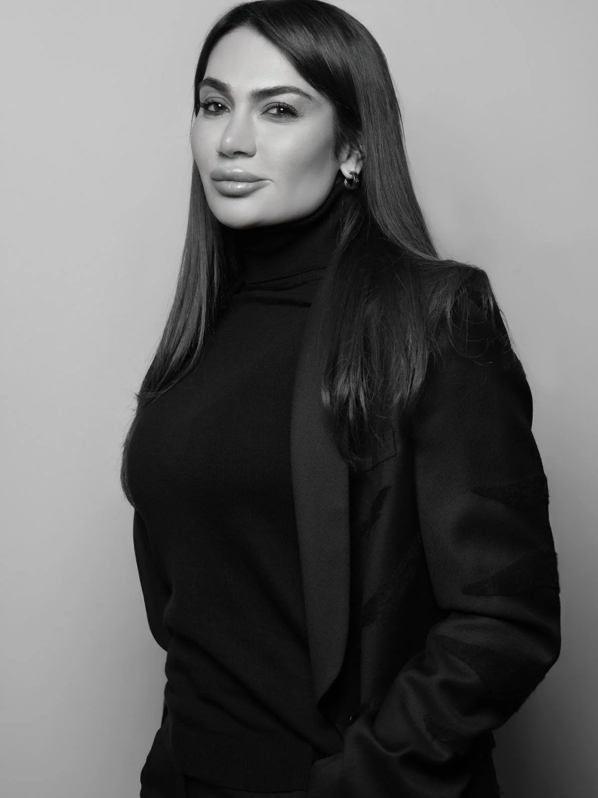 A portrait of a woman with long dark hair, wearing a black turtleneck and blazer, looking at the camera with a slight smile, in black and white.