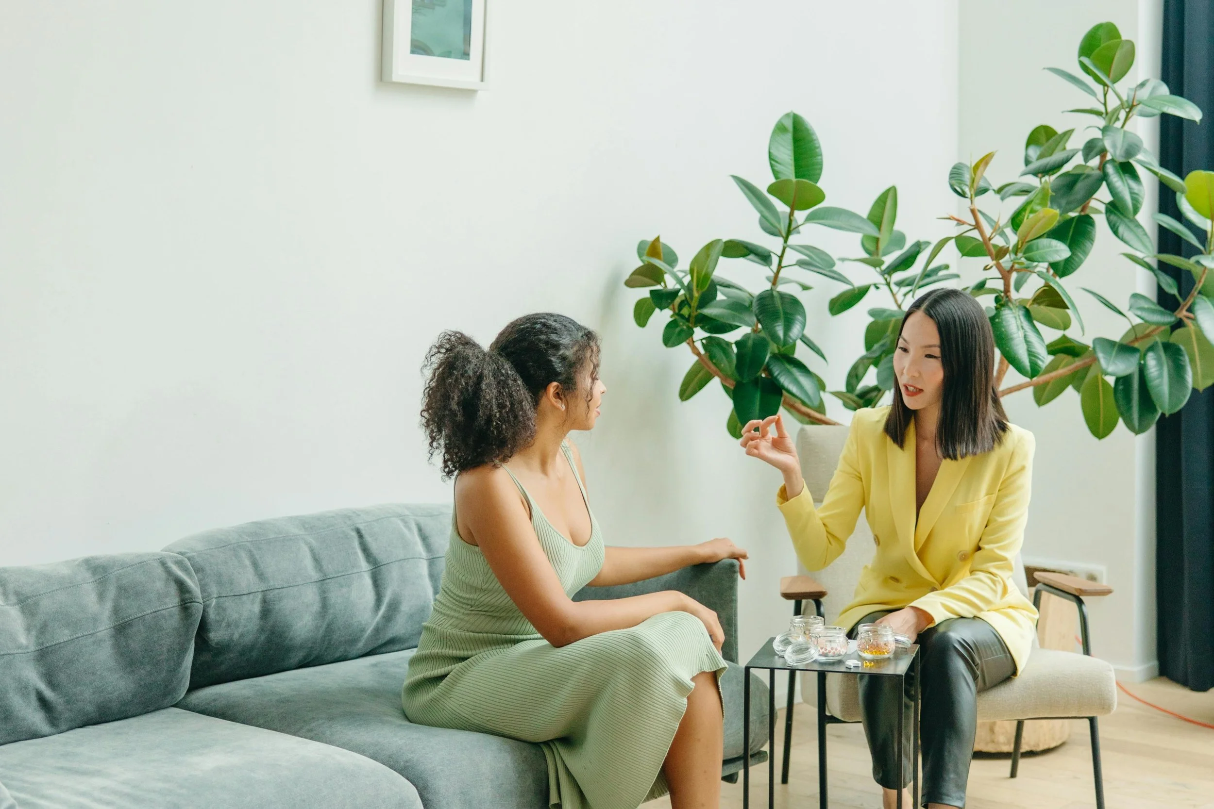 A woman with curly hair and a green dress sitting on a gray couch speaks with a woman in a yellow blazer and black pants who is seated on a gray chair. They are in a bright room with large green houseplants and a minimalistic decor.