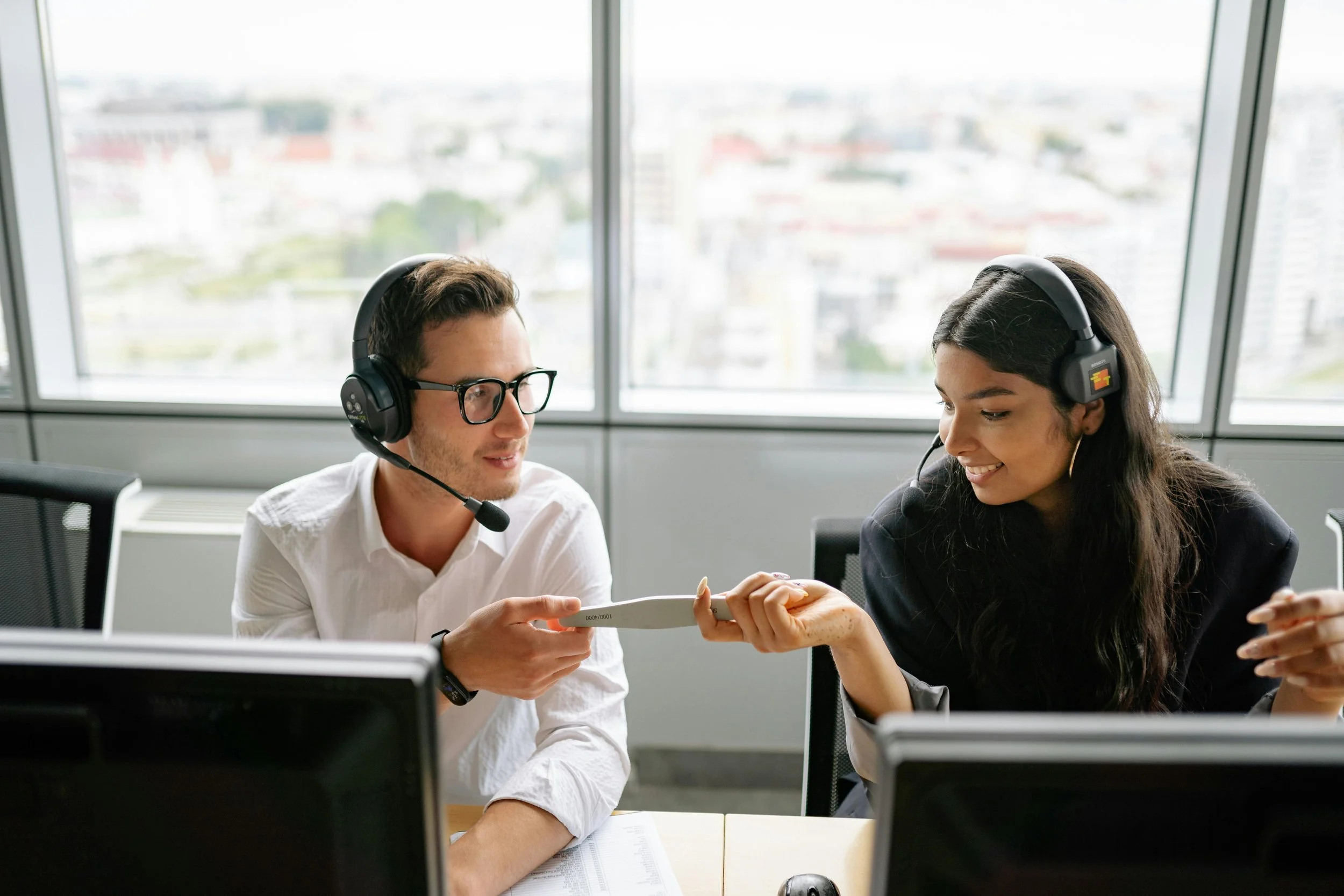 Two people in an office, wearing headsets, exchanging an object.