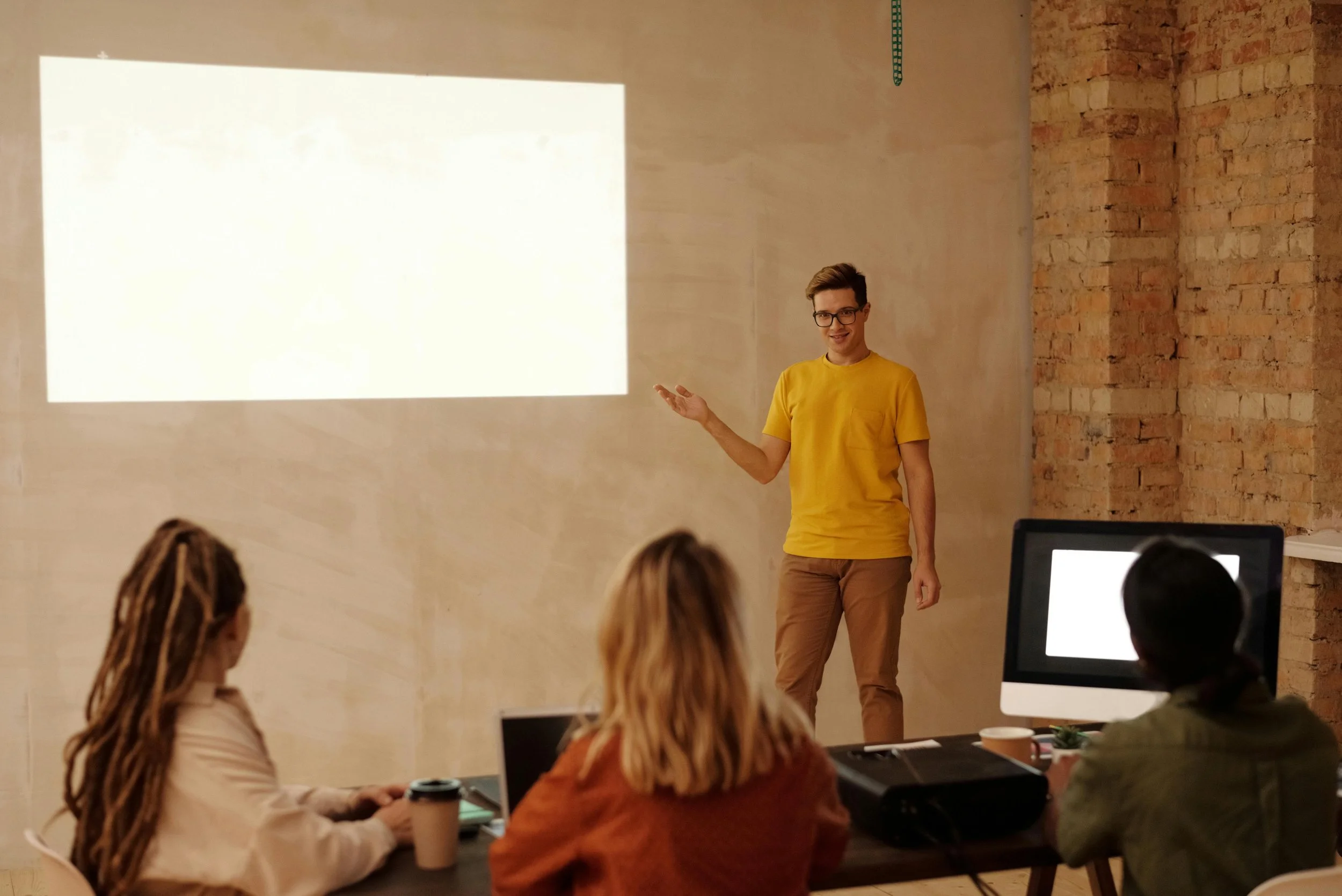 A young man with glasses and a yellow t-shirt is giving a presentation in front of an audience in a room with a brick wall and a projector screen.