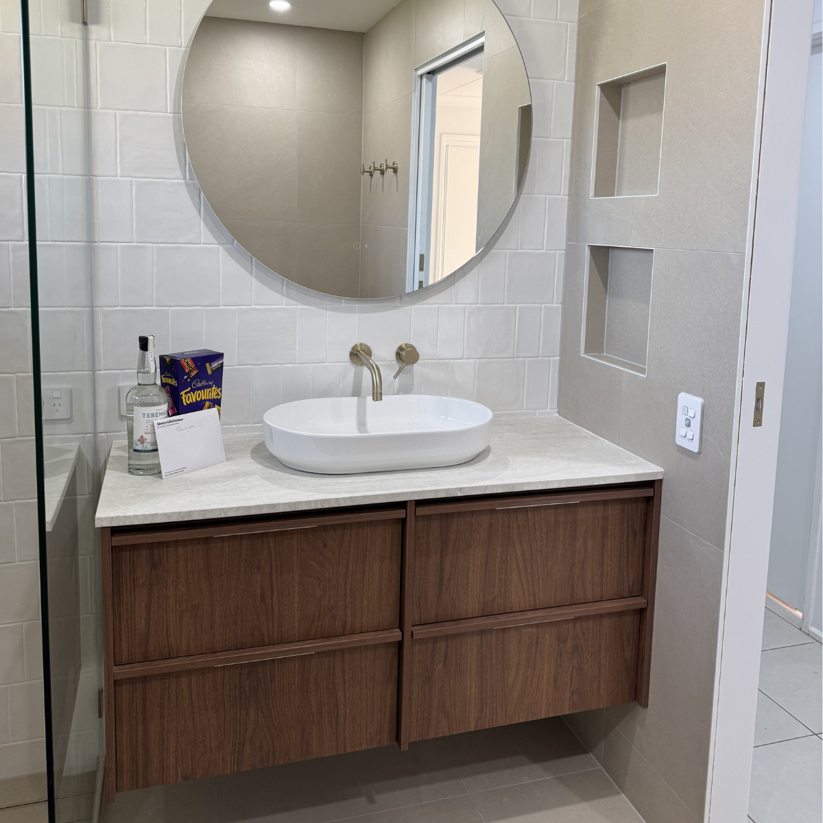 Bathroom vanity with a white vessel sink, a round mirror, and built-in wall shelves, with items like lotion, a box of snack, and a card on the countertop.