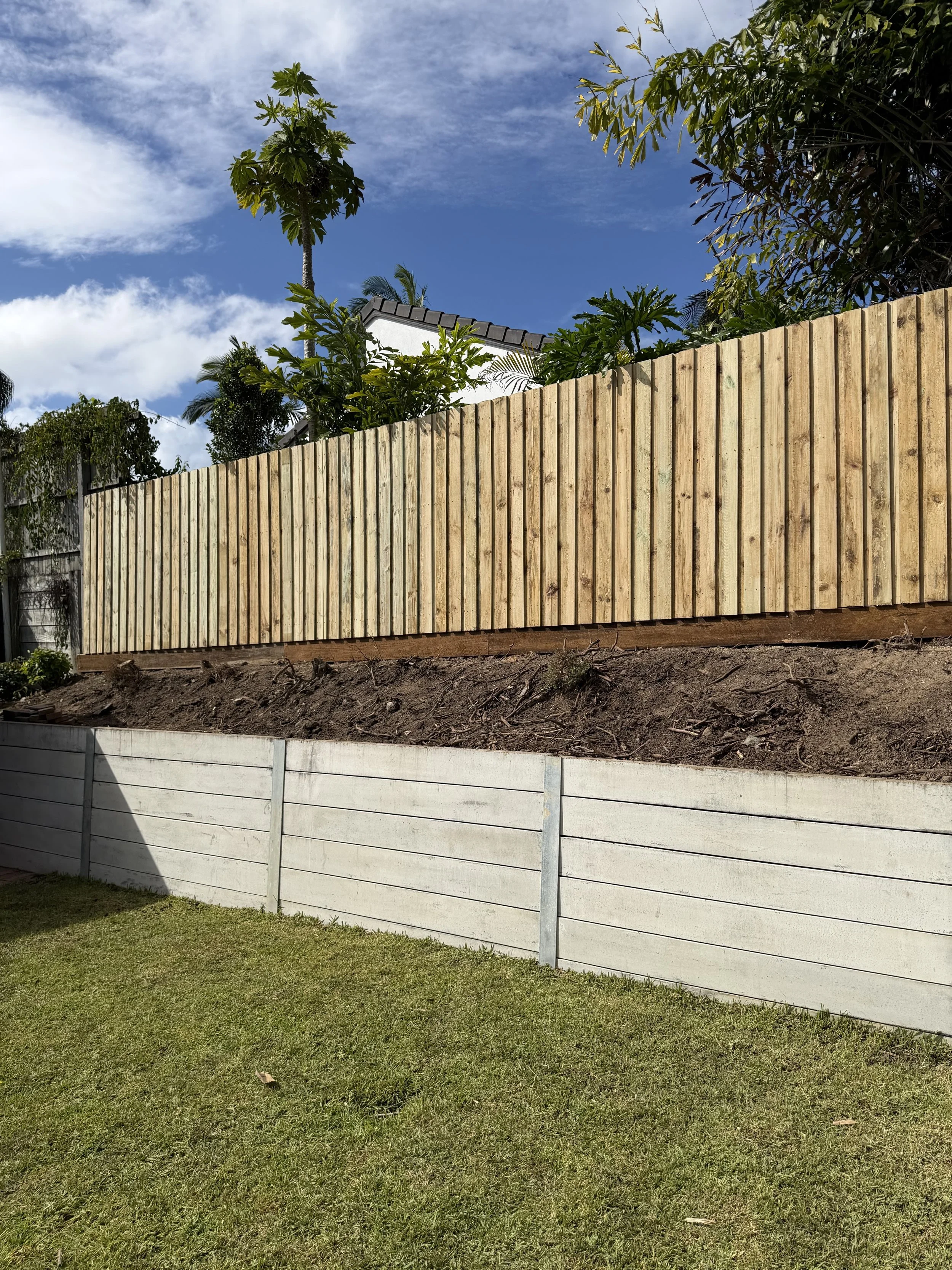 A beige metal fence along the side of a house with a grey tiled roof, backed by tall trees and a clear blue sky.