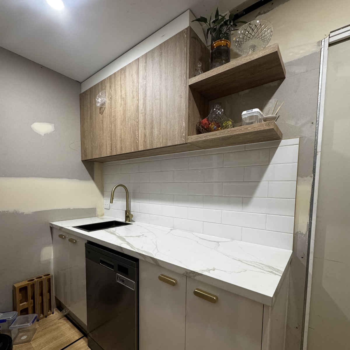 A small kitchen with white cabinets, marble countertop, gold handles, white subway tile backsplash, a black sink with a gold faucet, and upper wooden shelves holding jars and bowls. There is a dishwasher below the counter.