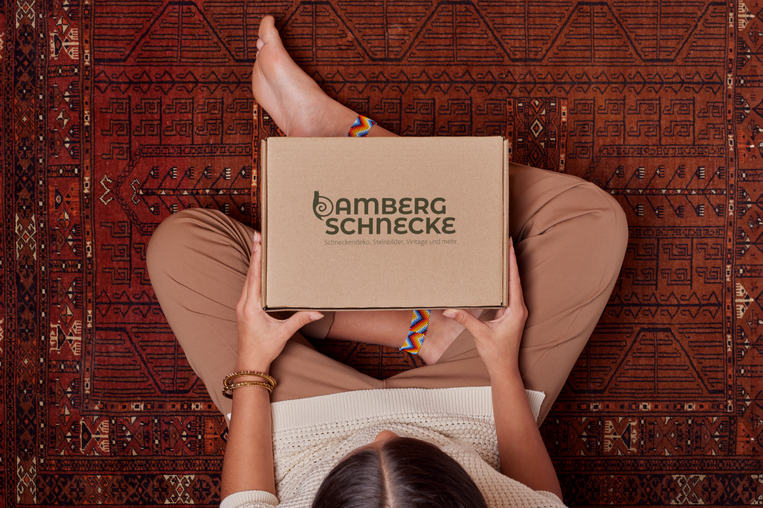Ein Mensch sitzt auf einem roten, orientalisch verzierten Teppich und hält eine braune Pappschachtel mit dem Aufdruck 'Amberg Schnecke' in den Händen, während die Person einen farbenfrohen Armband trägt.