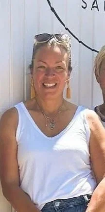 Smiling woman wearing sunglasses on head, white sleeveless top, and earrings standing outdoors with a white fence in the background.