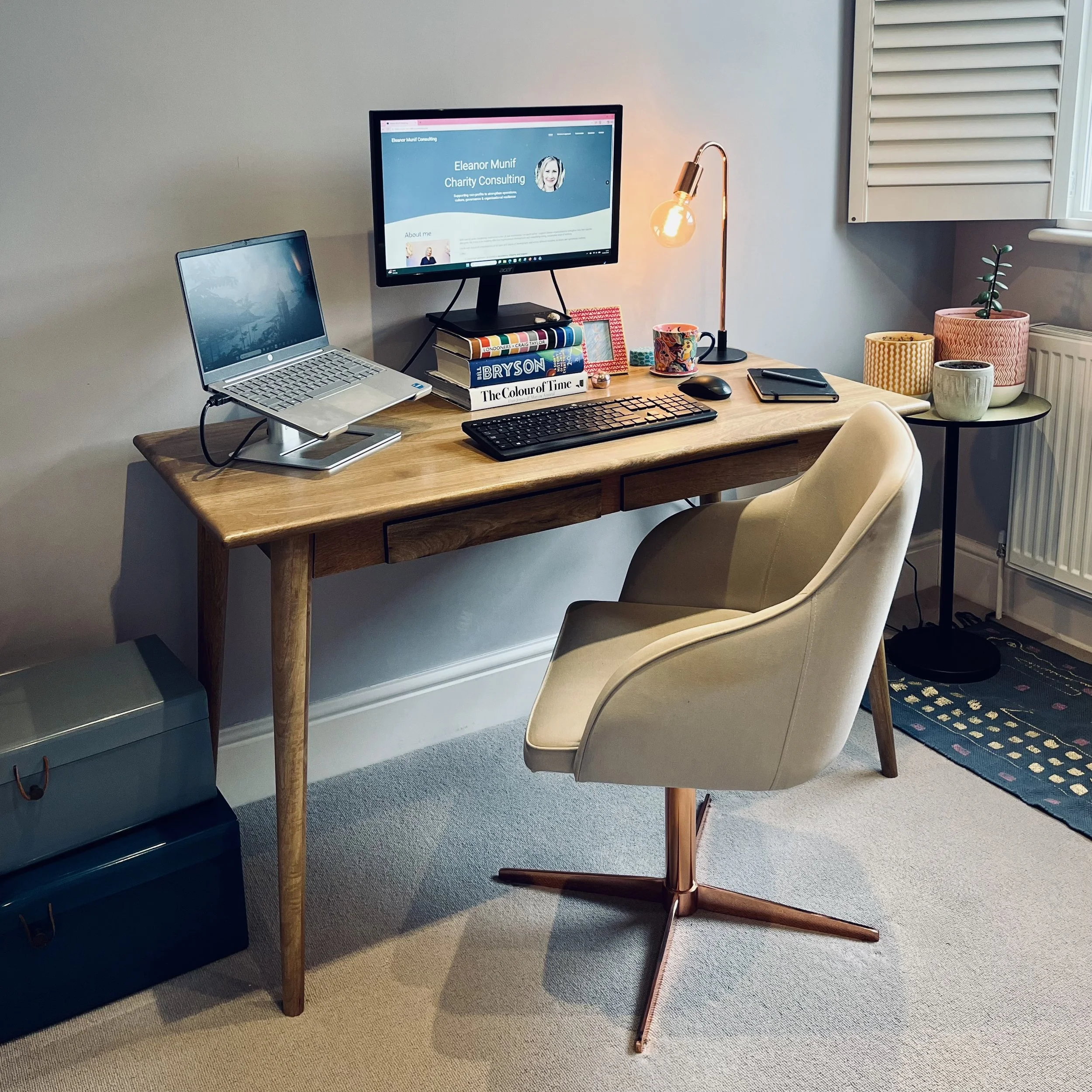 Home office desk with a laptop on a stand, a second monitor displaying a personal website, a stack of books, a keyboard, a mouse, a notebook, a smartphone, a table lamp, and decorative planters with plants nearby.