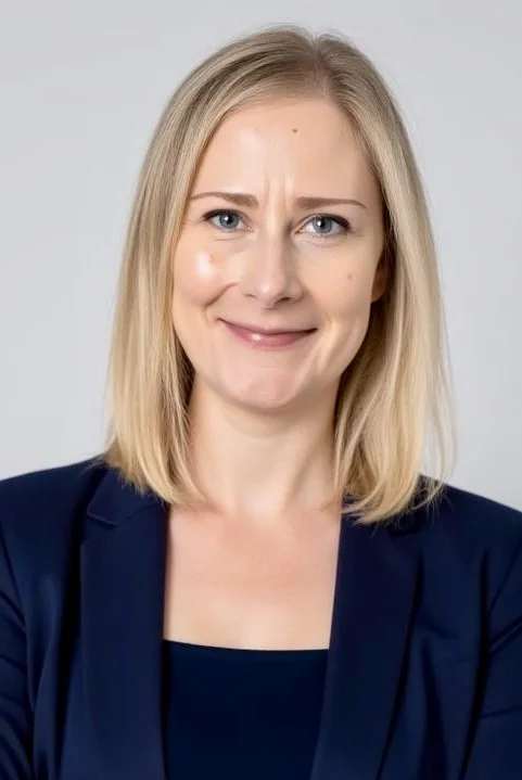 A woman with blonde hair and a slight smile, wearing a navy blazer and navy top, standing against a light gray background.