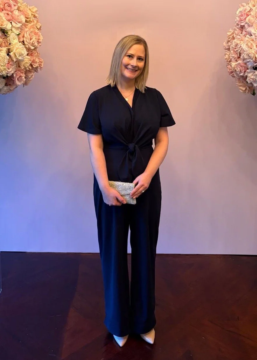 A woman in a black jumpsuit holding a silver clutch, standing in front of a pink wall decorated with large pink and cream hydrangea flower arrangements, smiling at the camera.