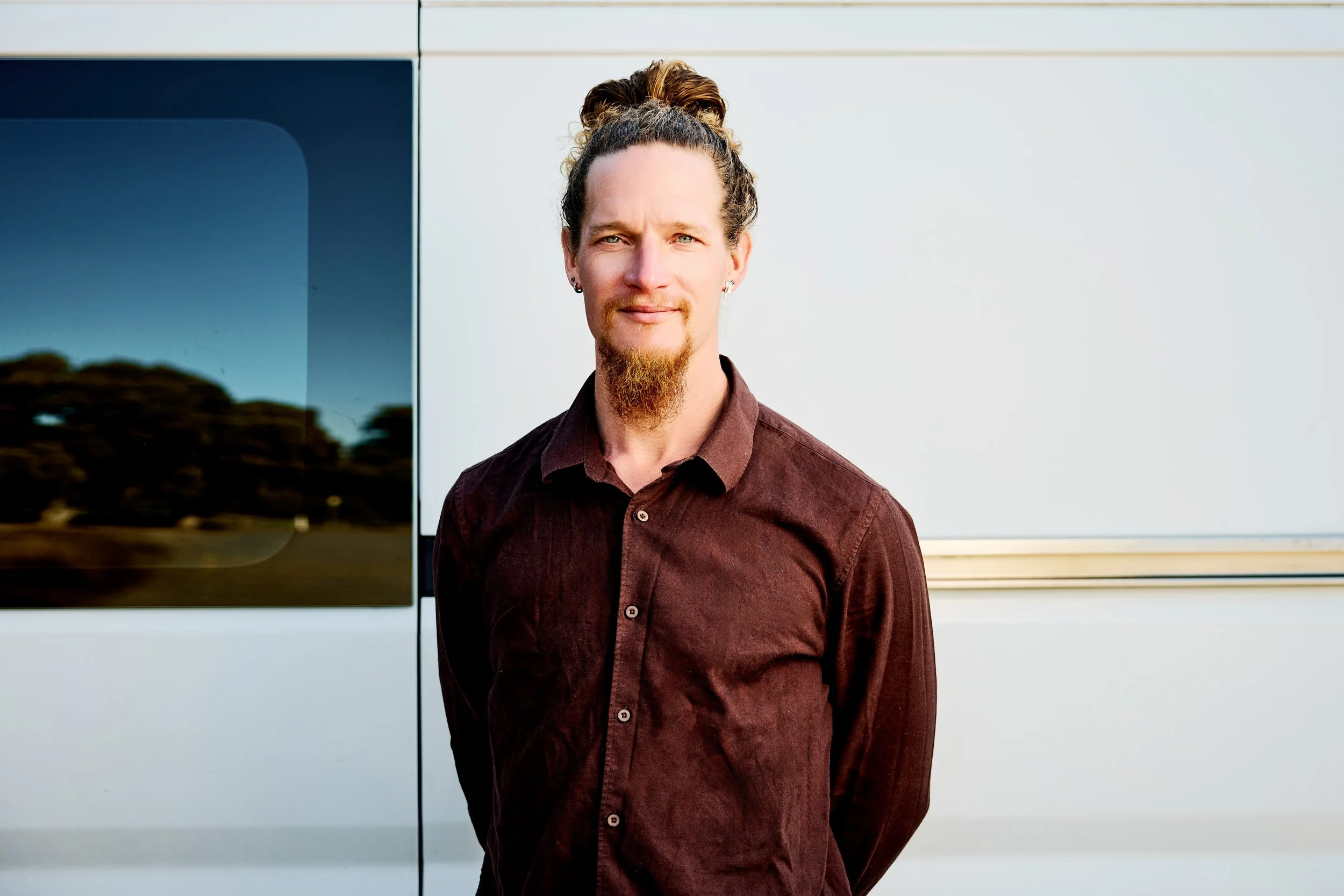 Portrait of a man with curly hair tied in a bun, a beard, and earrings, standing outdoors in front of a vehicle.