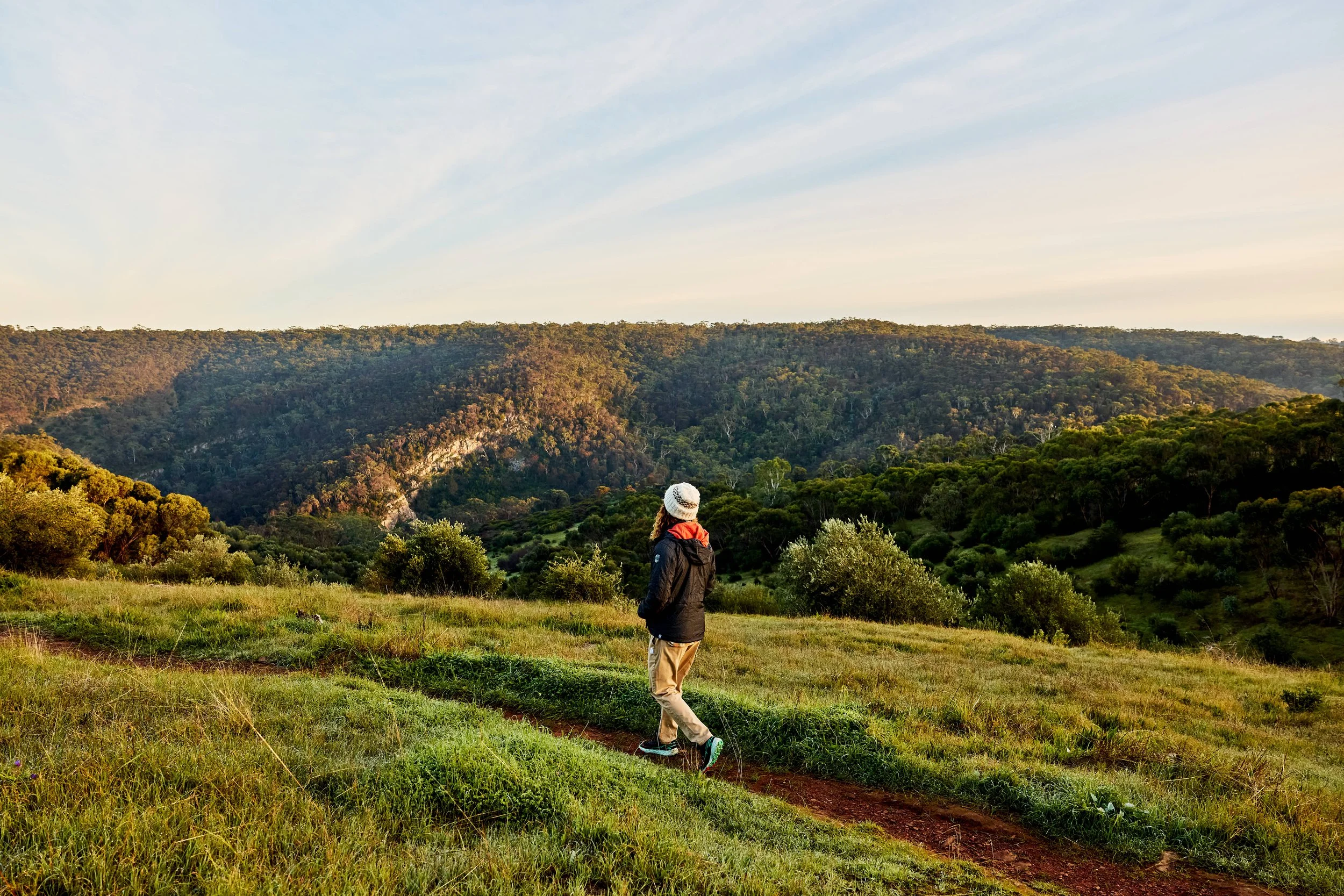 A person walking along a dirt trail in a grassy field with rolling hills and dense forest in the background under a partly cloudy sky.