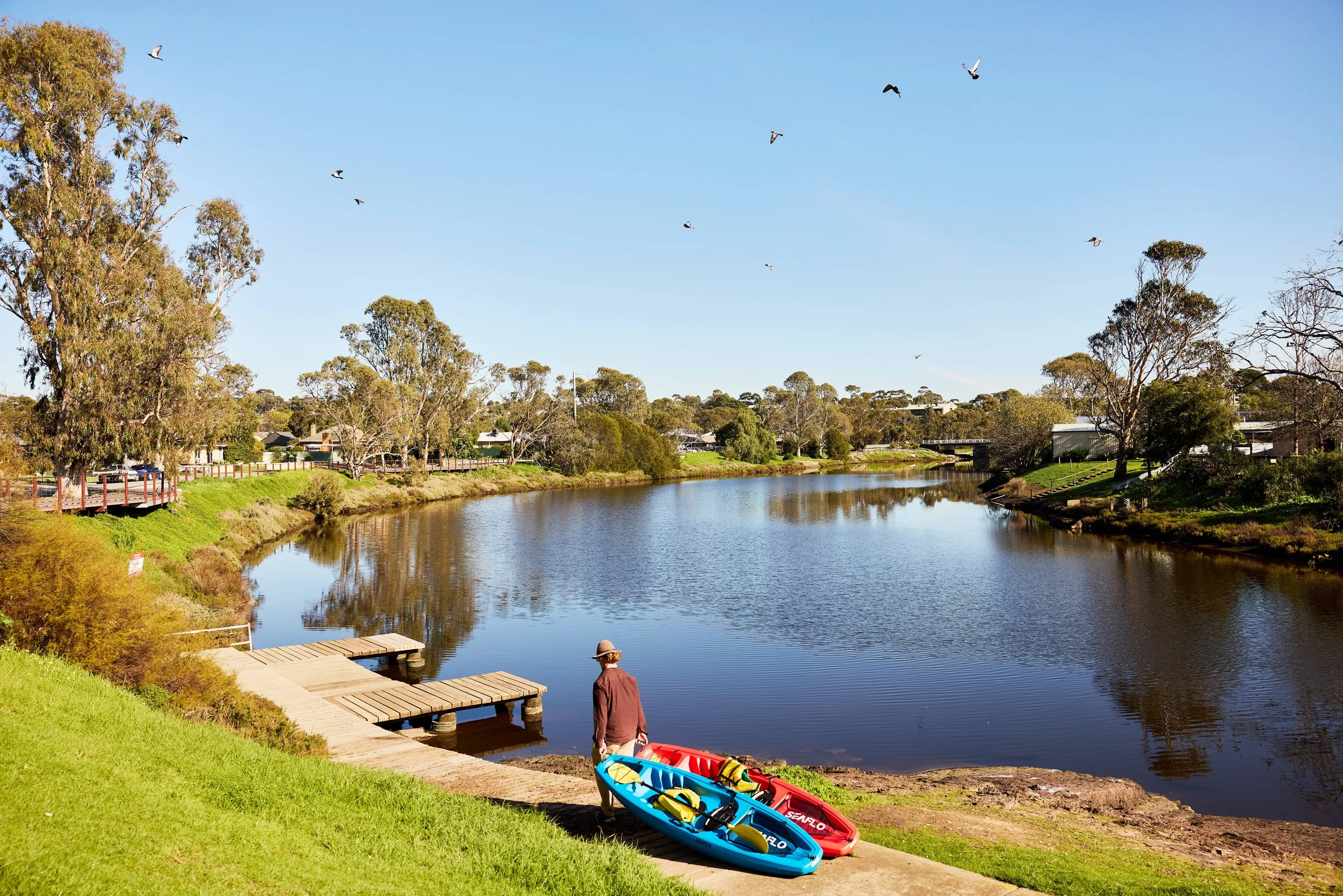 A peaceful river scene on a sunny day with a man standing near the water, two colorful kayaks on the shore, and birds flying in the clear blue sky among tall trees and residential houses.