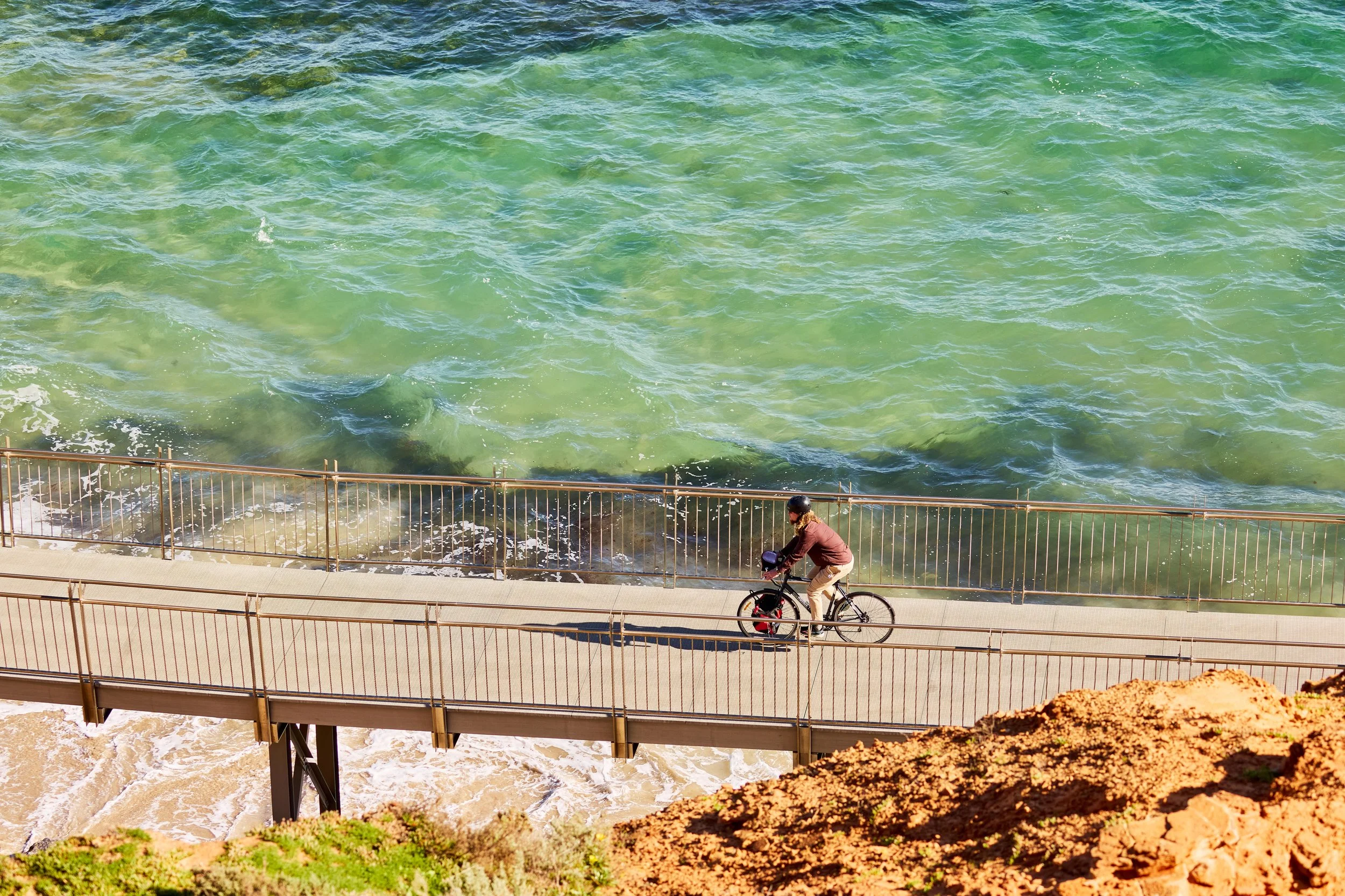 Person riding bicycle along a walkway by the ocean with waves crashing against rocks and a dirt hillside in the foreground.