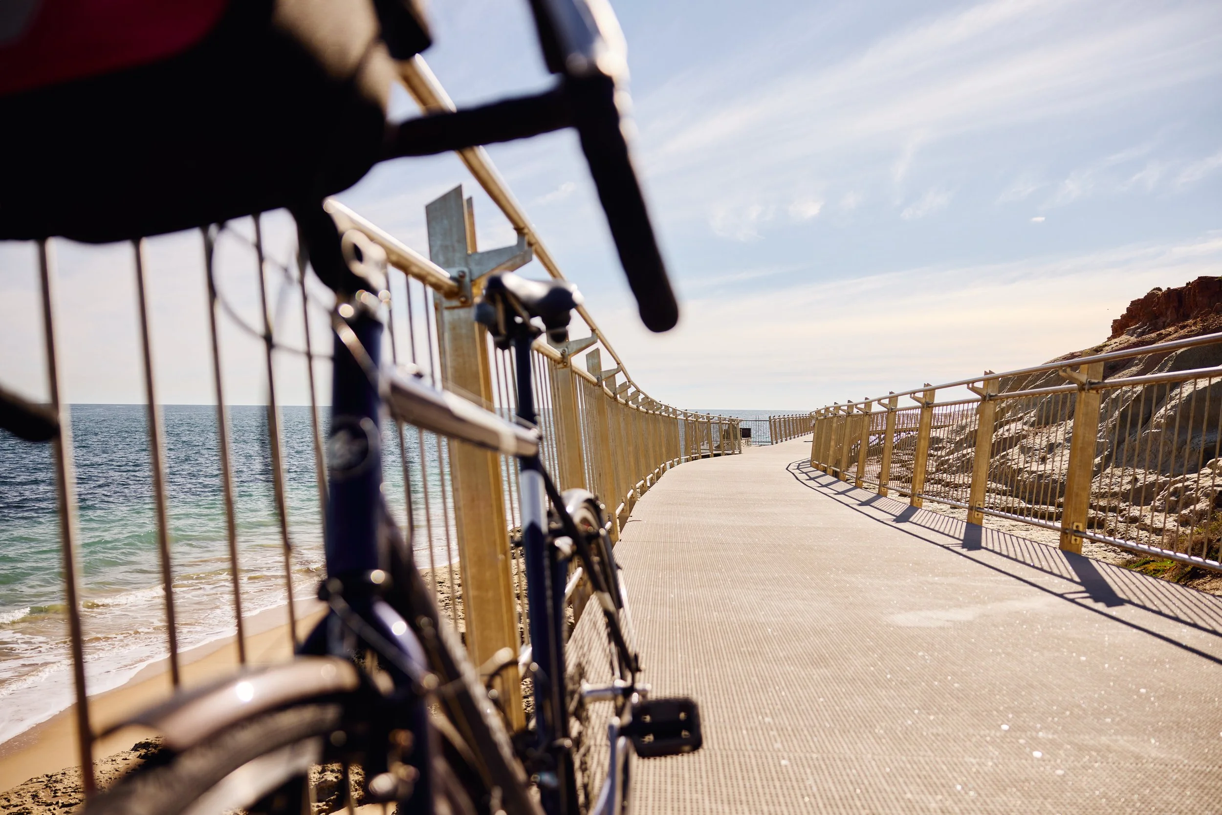 Bicycles on a railing next to a beach boardwalk with a view of the ocean and clear sky.