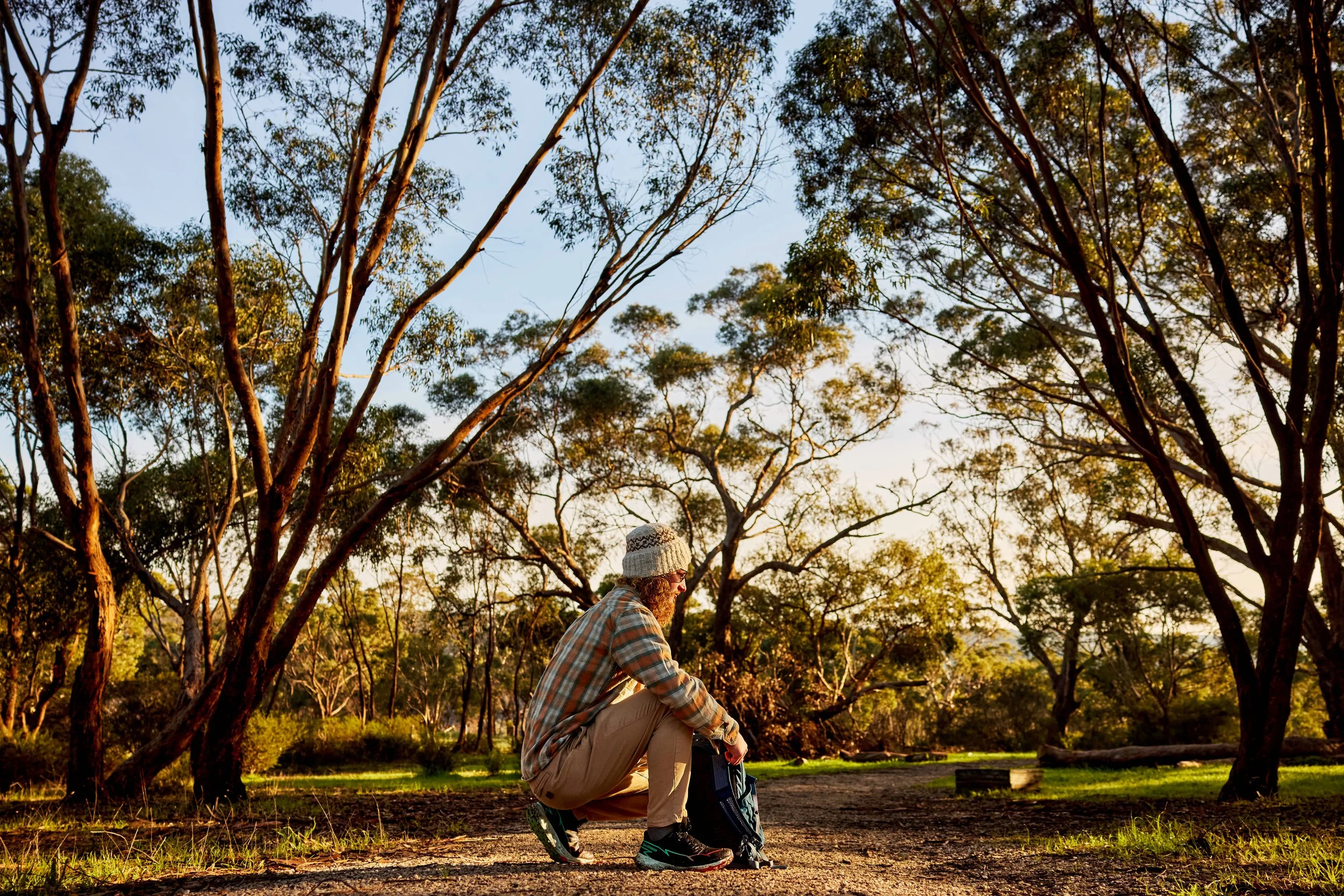 A person wearing a plaid shirt, khaki pants, and a hat is packing his backpack for a walk in a forested park during sunrise, with tall trees and blue sky in the background.