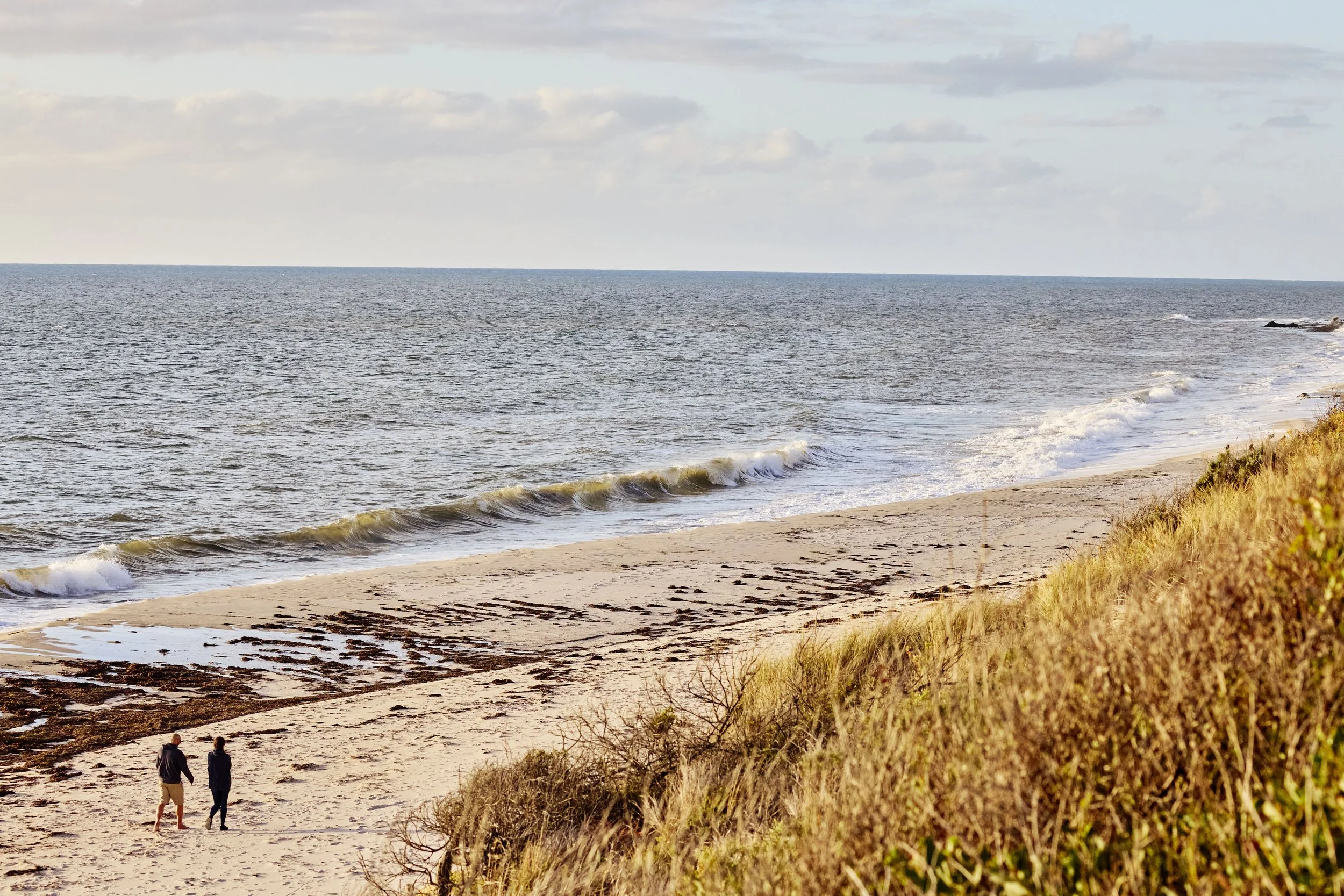Two people walking along a sandy beach with waves and grassy dunes under a cloudy sky.