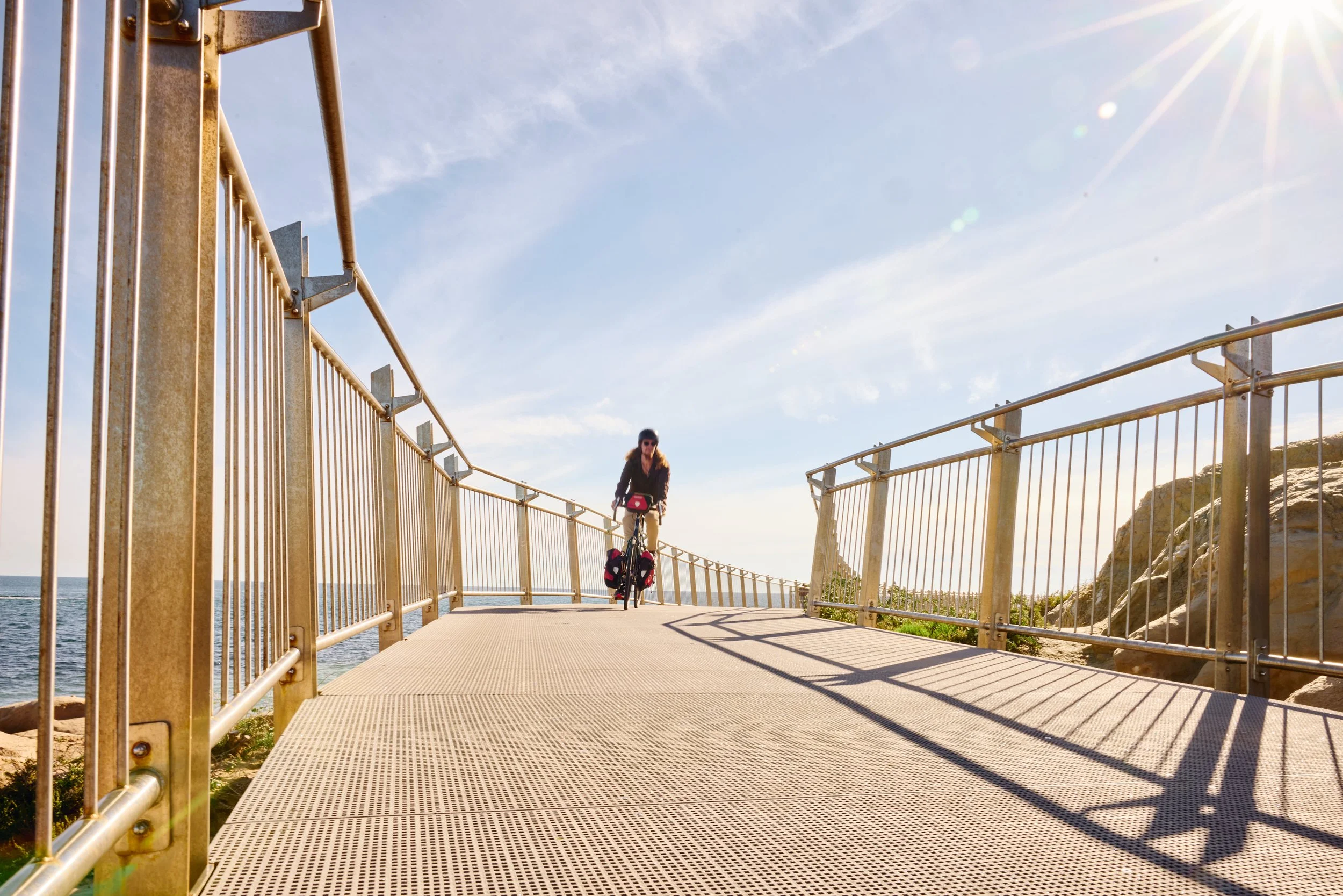 A person riding a bicycle on a board walk with railings, near the beach under a clear blue sky with the sun shining brightly.