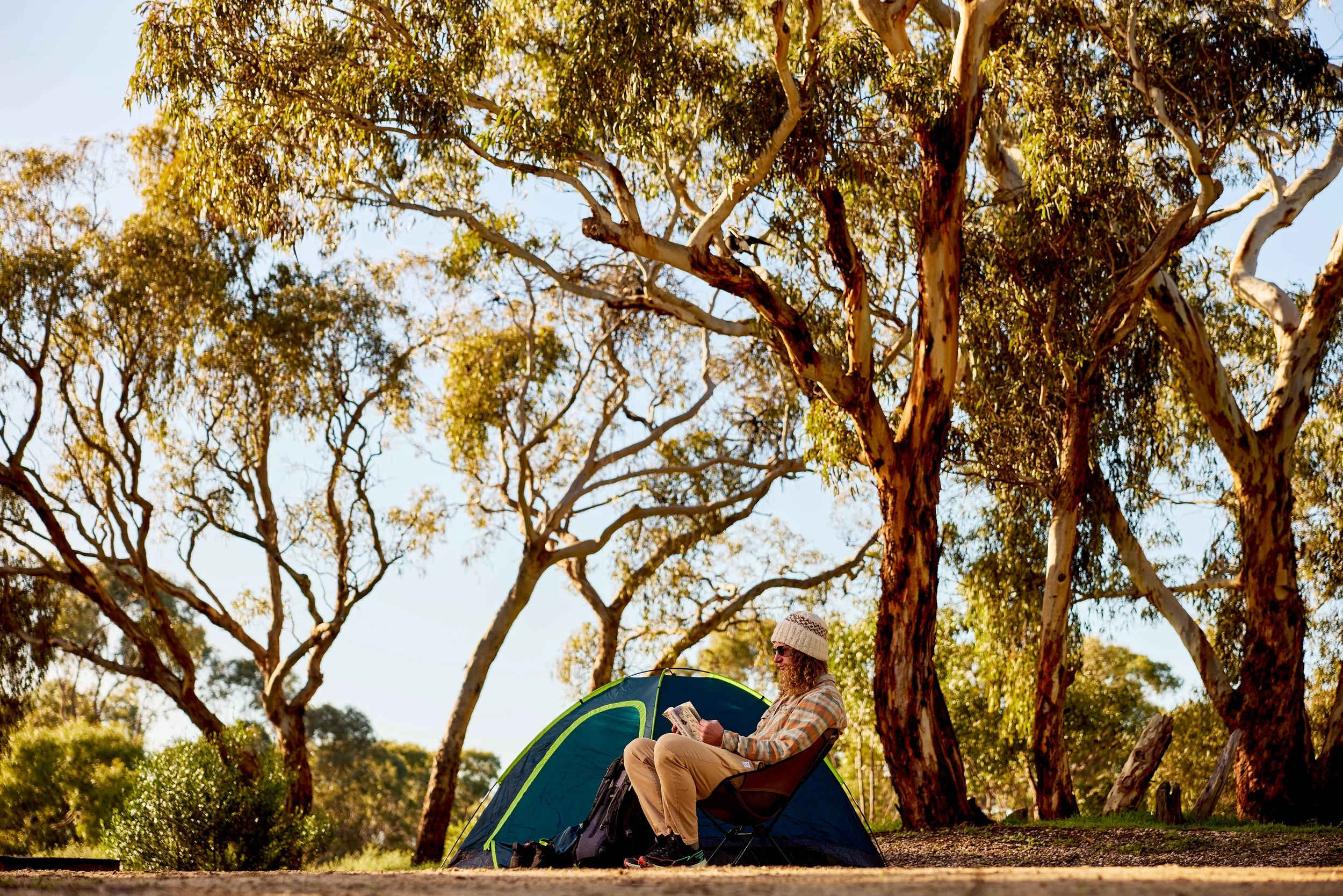 A person sitting in a camping chair outside near a small tent, reading a book or magazine, with large trees and blue sky in the background.