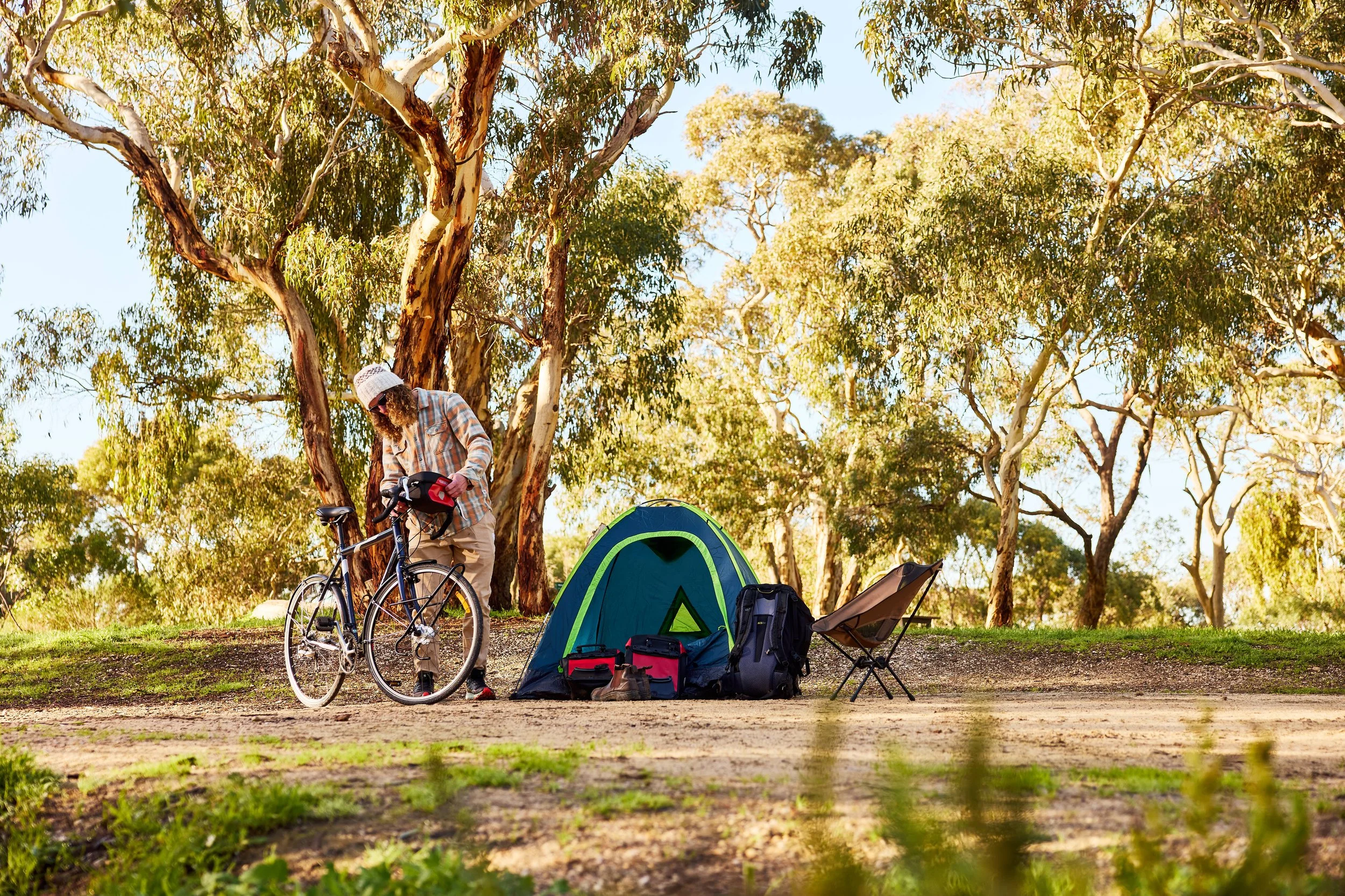 A person in a plaid shirt and white hat preparing a bicycle next to a green and blue tent in a wooded campsite with backpacks, chairs, and camping gear, under tall trees with green leaves.