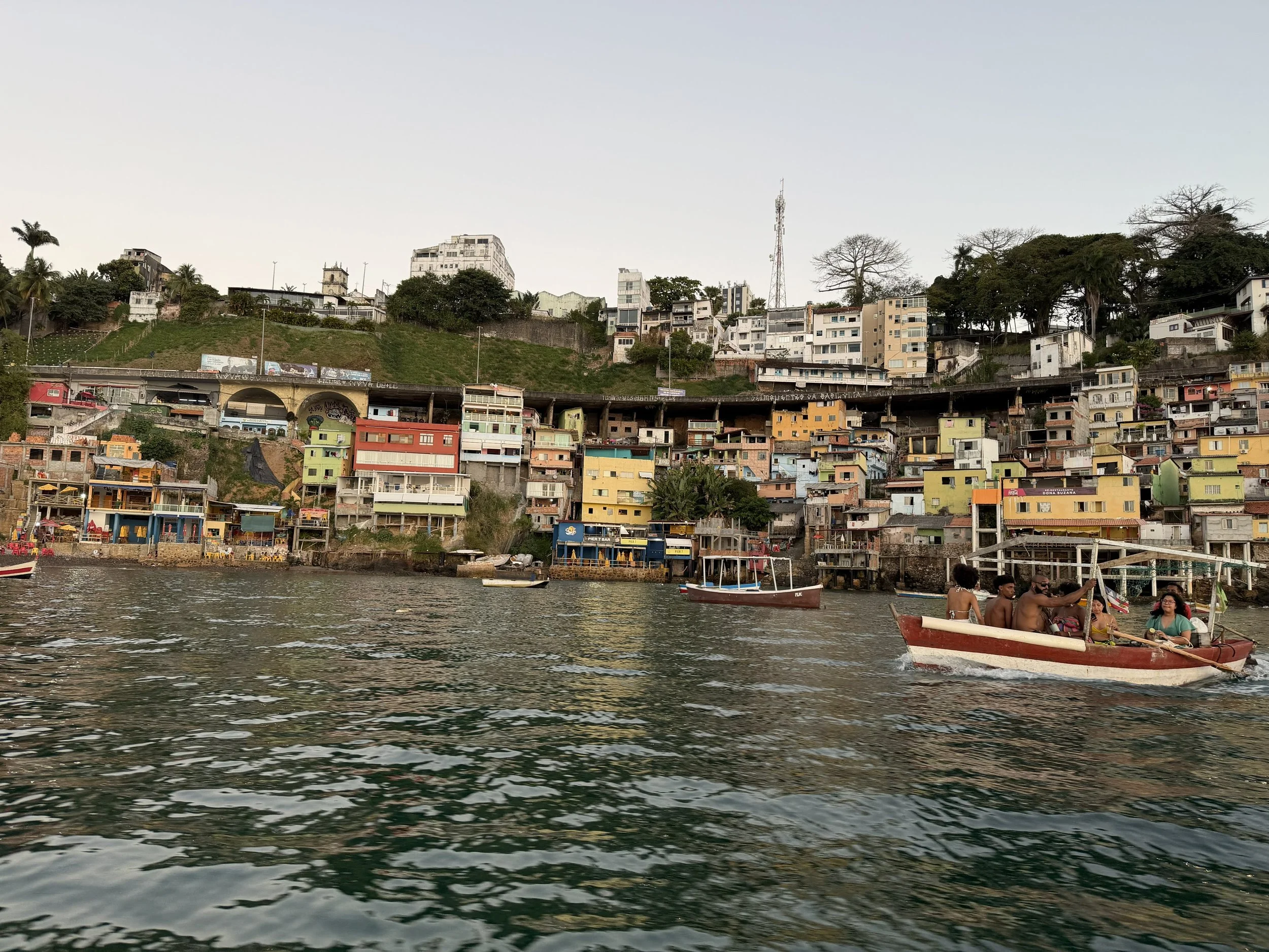 Waterfront view of a hillside neighborhood with colorful houses and trees, and a boat with people in the foreground.