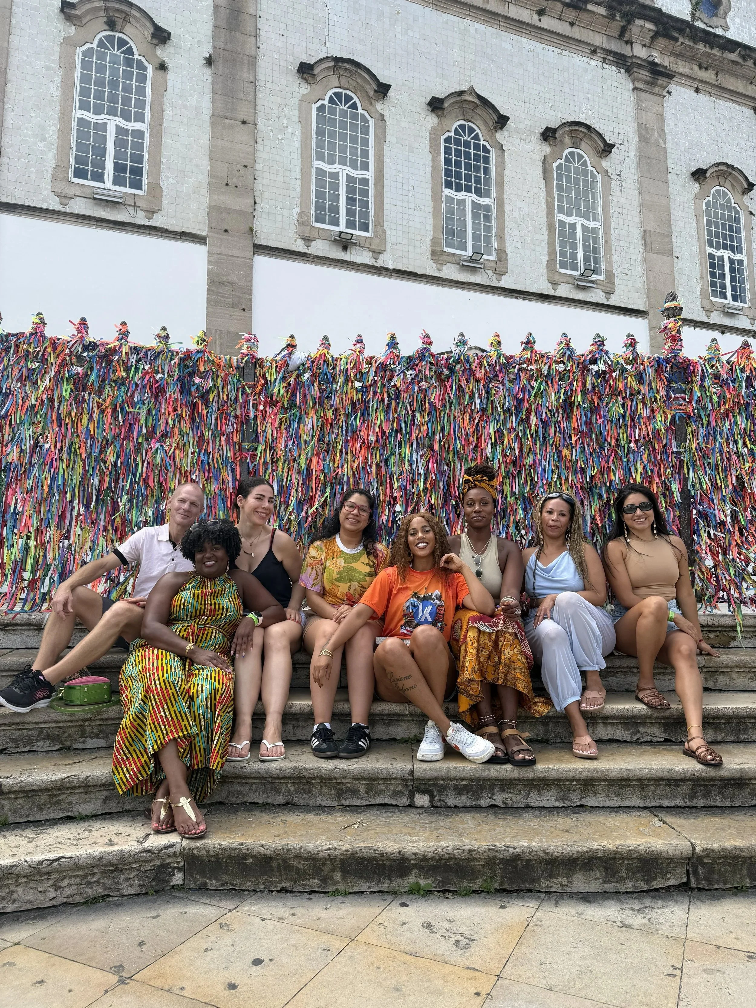 Group of diverse individuals sitting on stone steps in front of a decorative wall covered with colorful ribbons, with an old building featuring large arched windows in the background.