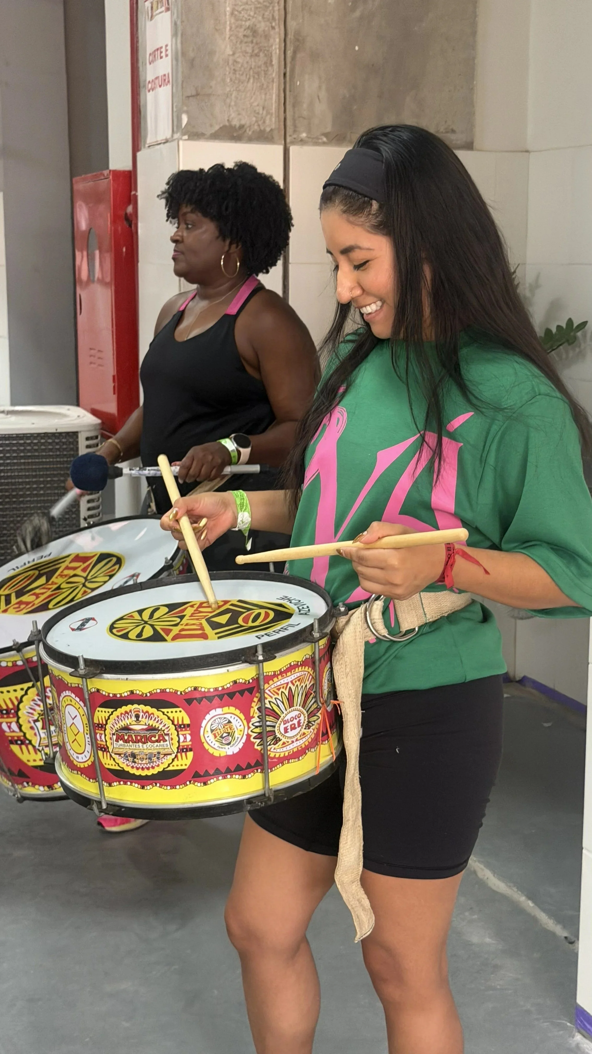 A young woman smiling and playing a percussion drum with drumsticks, wearing a green t-shirt and black shorts, indoors at a festival or music event.