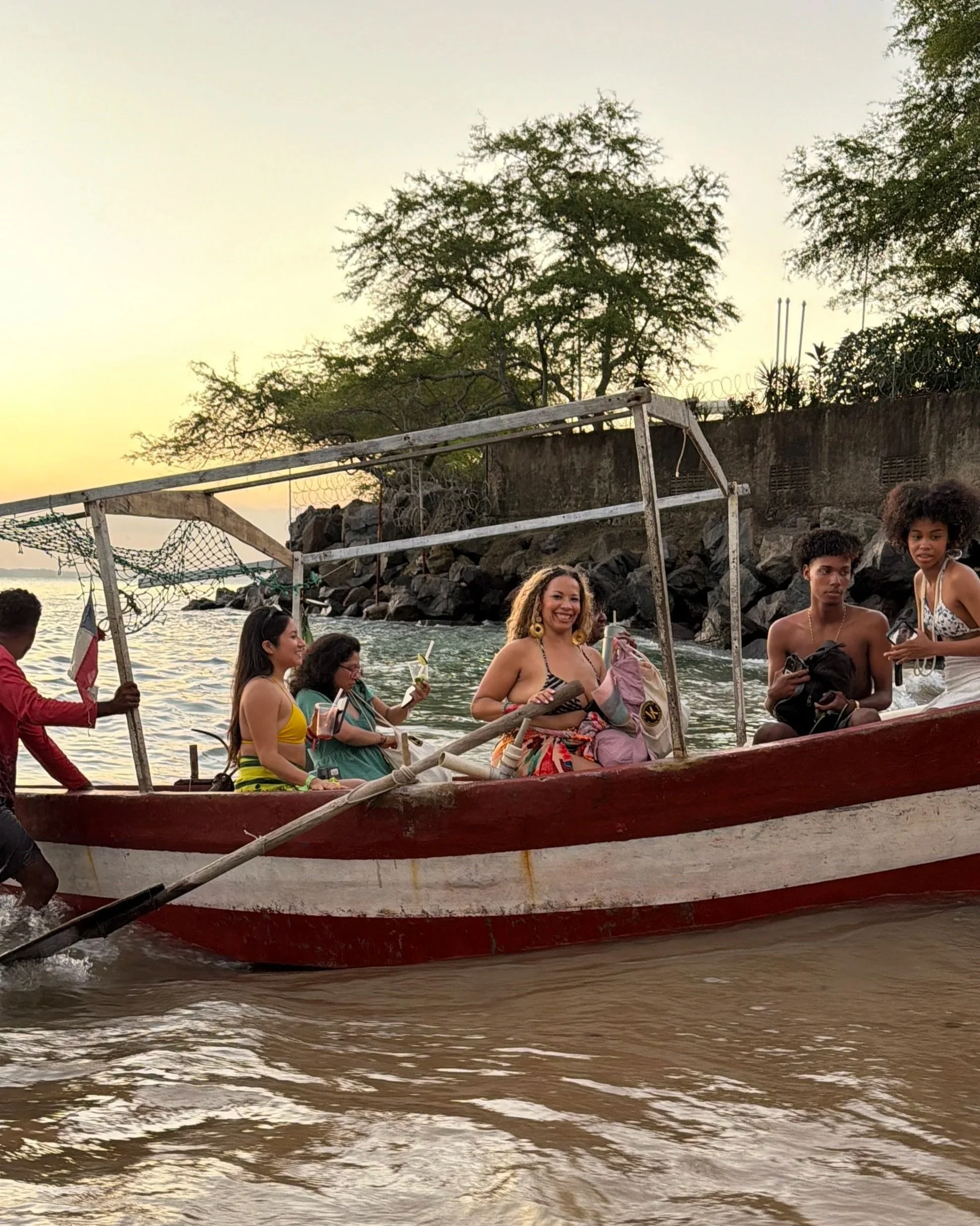 People on a boat in the water during sunset, with trees and a stone wall in the background.