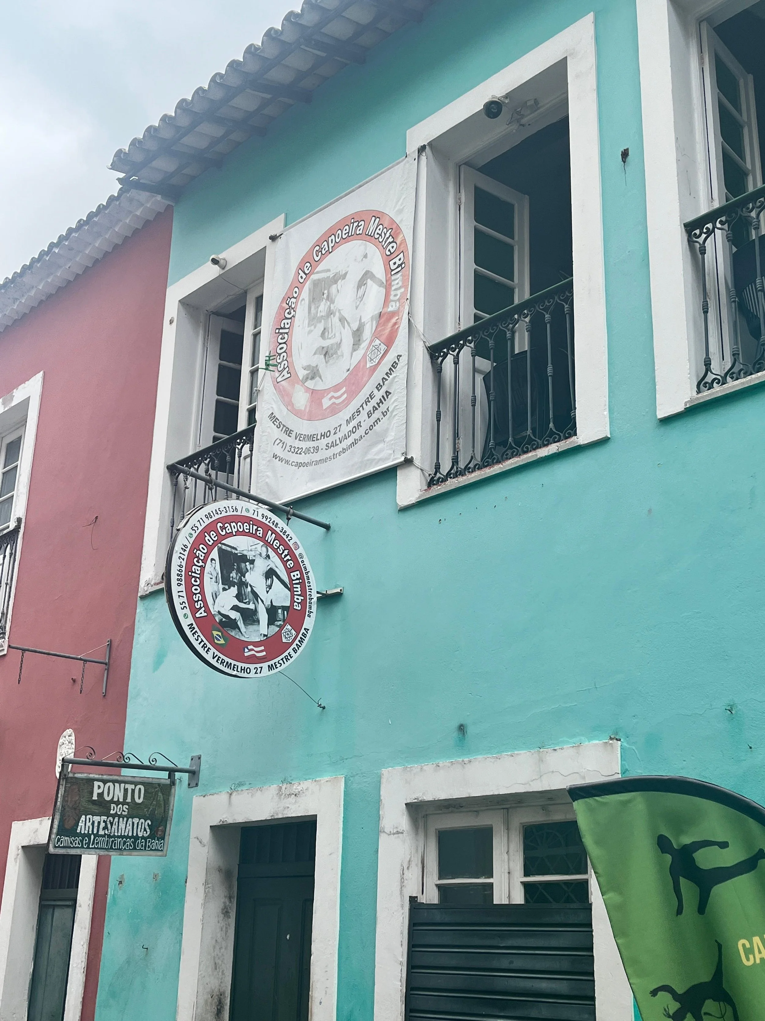 Colorful building with a light blue and pink facade, featuring signs for a local art and craft shop and a martial arts school called Associação de Capoeira Mestre Bimba, with open windows and a small balcony.