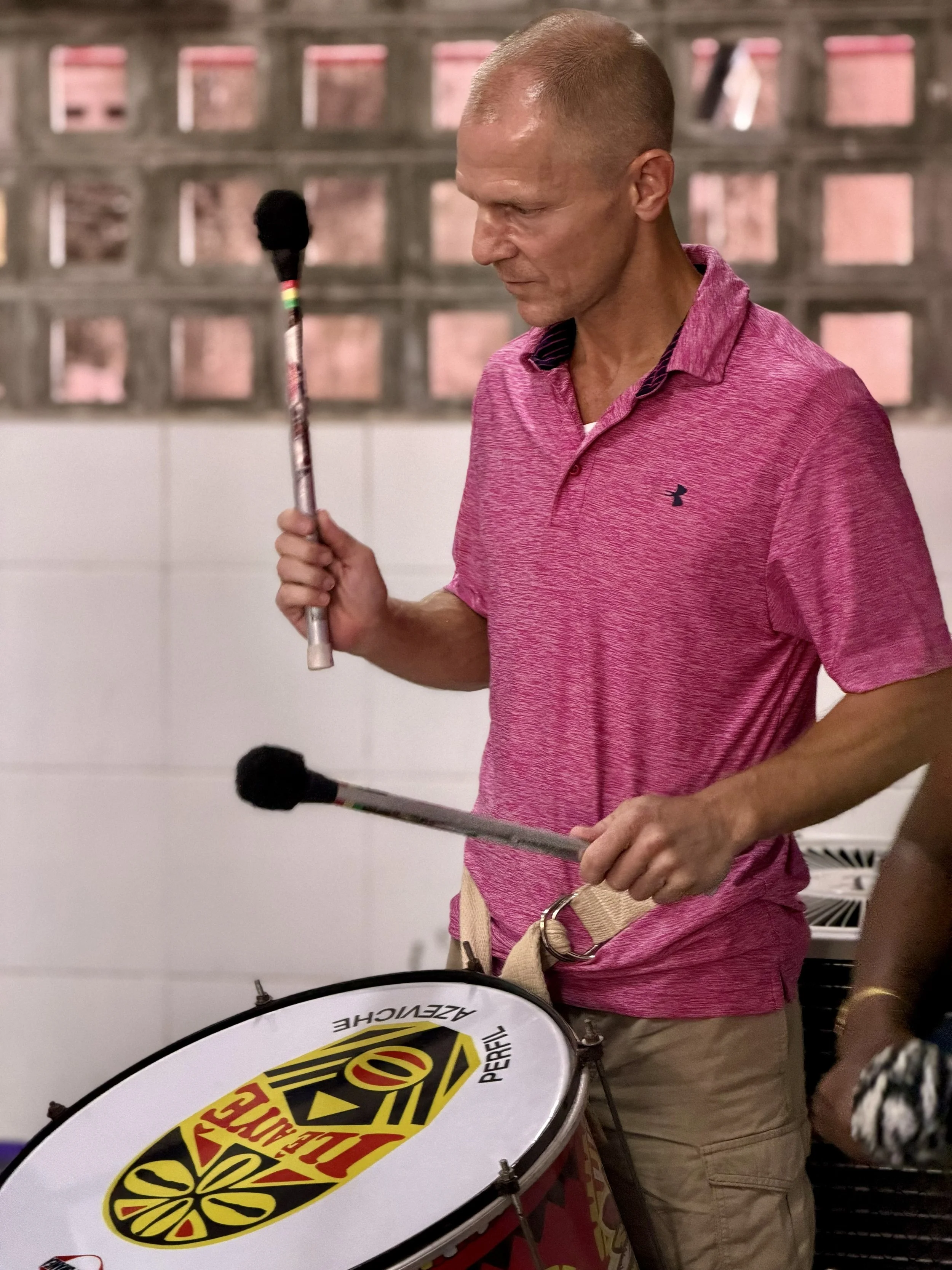 A man in a pink shirt holding a drumstick with the logo of a pizza brand, standing near a drum with the same logo, in an indoor setting.