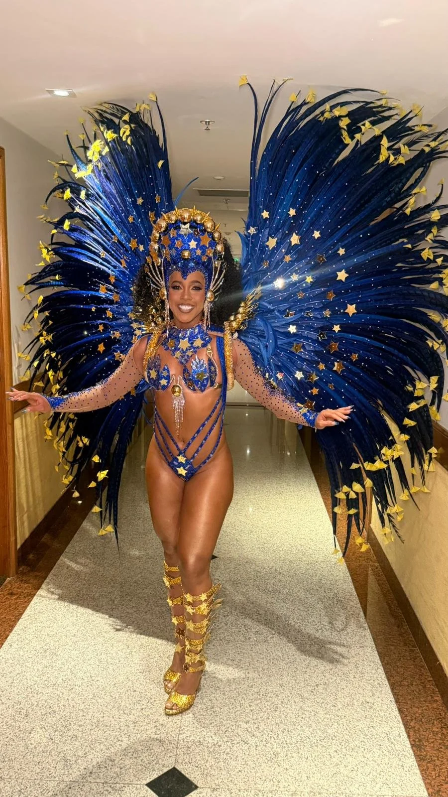 A woman dressed in a vibrant, elaborate costume with large blue and yellow feathered wings adorned with stars, standing in a hallway.