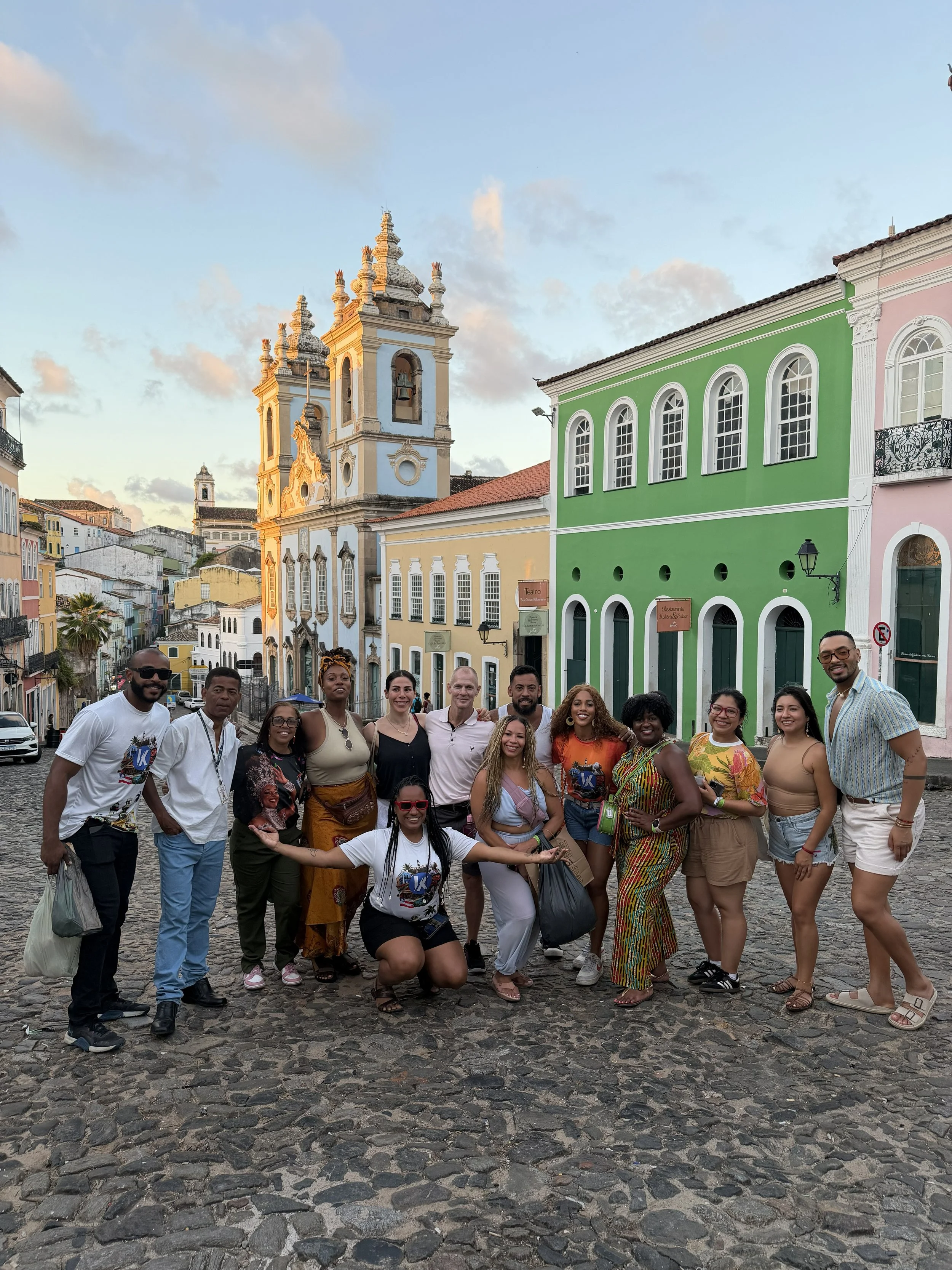 A group of diverse people smiling and posing on a cobblestone street in front of colorful colonial-style buildings and church towers during the daytime.