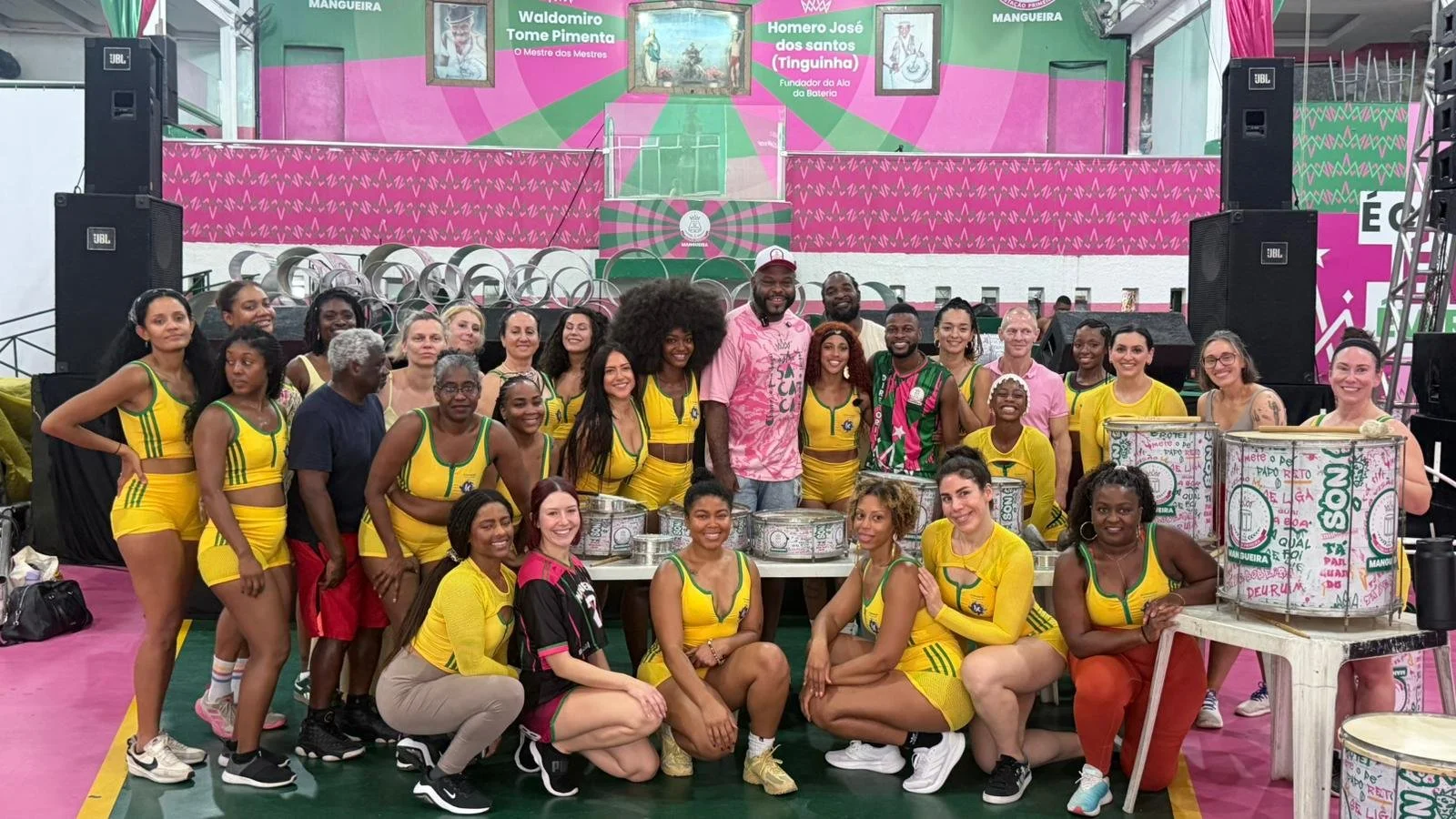 Group of women and men celebrating in a colorful indoor space with a large pink, green, and white mural in the background, some wearing yellow sports uniforms and others in casual clothing, with drums and musical equipment nearby.