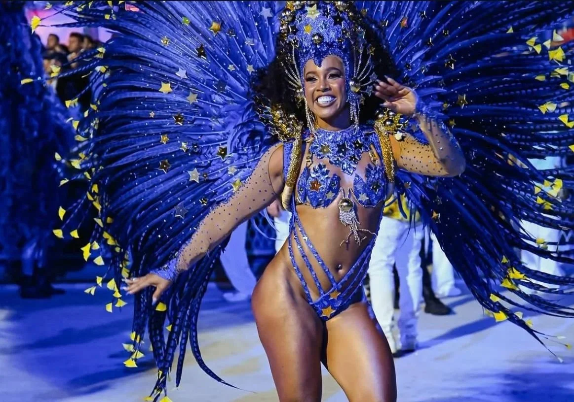 A woman in a vibrant, elaborate carnival costume with blue feathers and gold accents, smiling and waving.