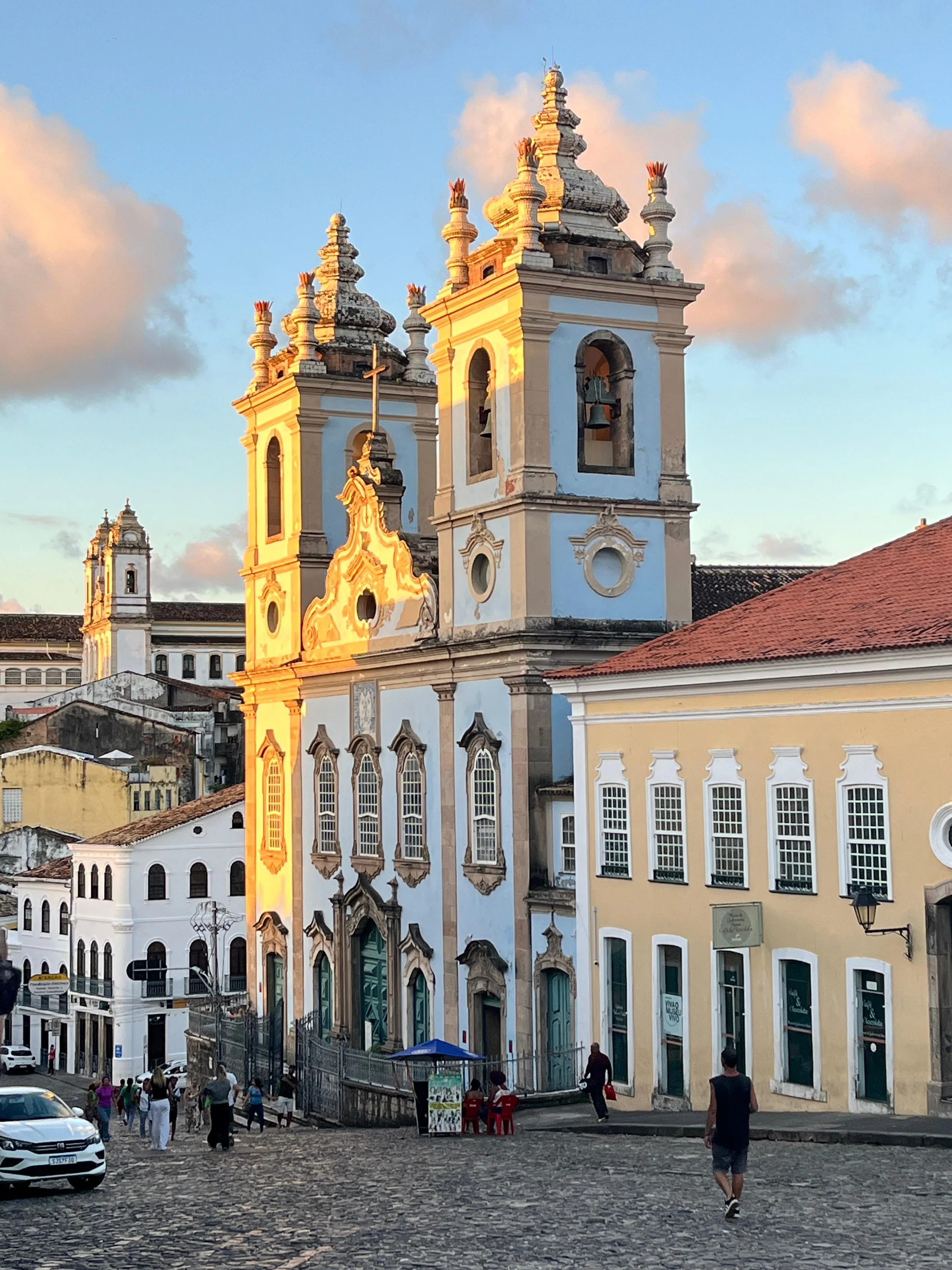 Historical church with two bell towers in a cobblestone plaza during sunset, with pedestrians and cars in the foreground.