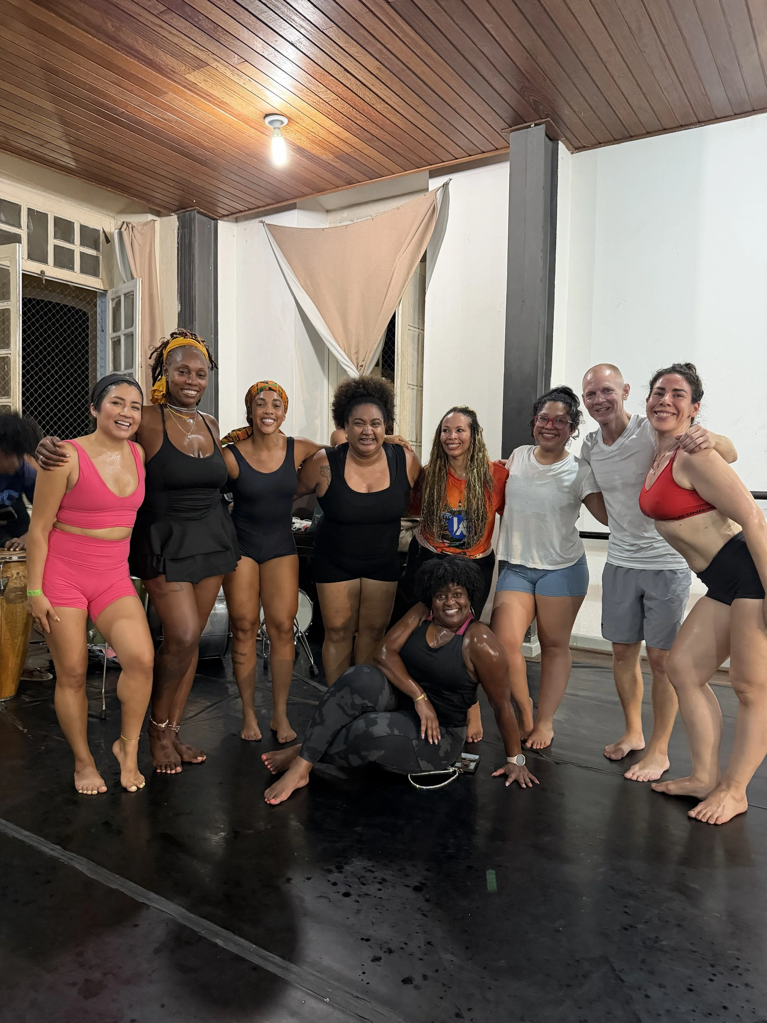 A group of diverse people smiling and posing together in a dance studio, some with arms around each other, with a wooden ceiling and a drum set in the background.