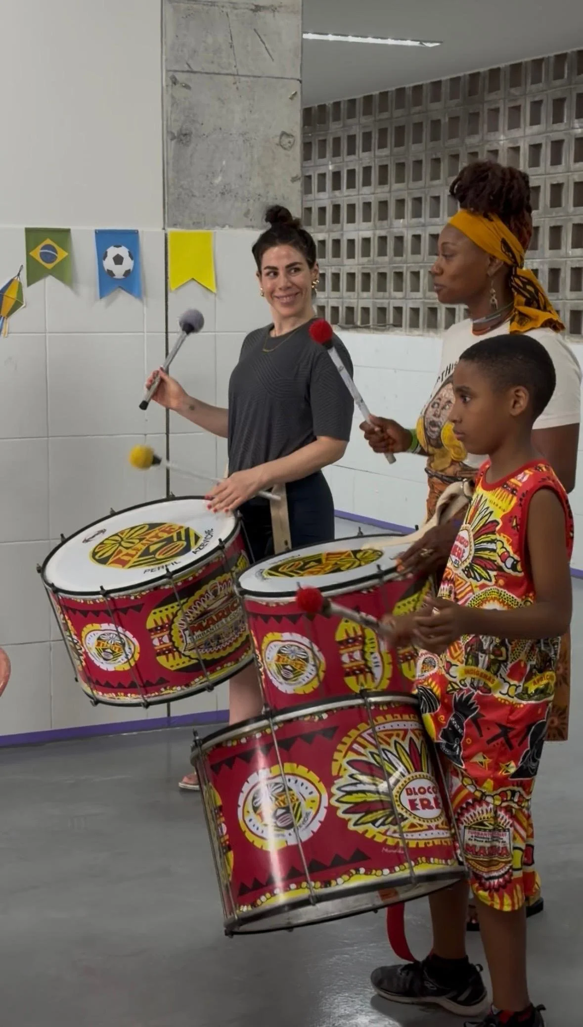 Two women and a boy playing drums; one woman has dark hair in a bun and is smiling, wearing a gray outfit. The other woman has braided hair with a yellow headscarf, wearing a white shirt with a graphic and colorful shorts. The boy has short hair and 