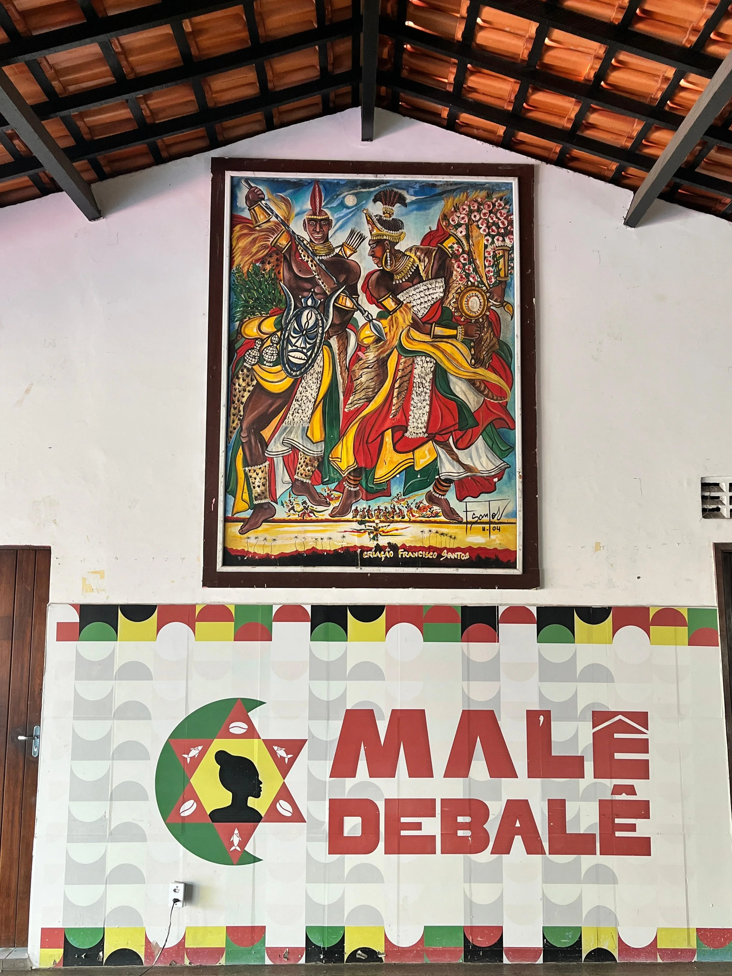 A colorful painting depicting two traditional African warriors in ceremonial attire, holding weapons, with a moonlit sky in the background. The painting is framed and hung on a white wall. Below the painting, there is a large sign with a logo, a silh