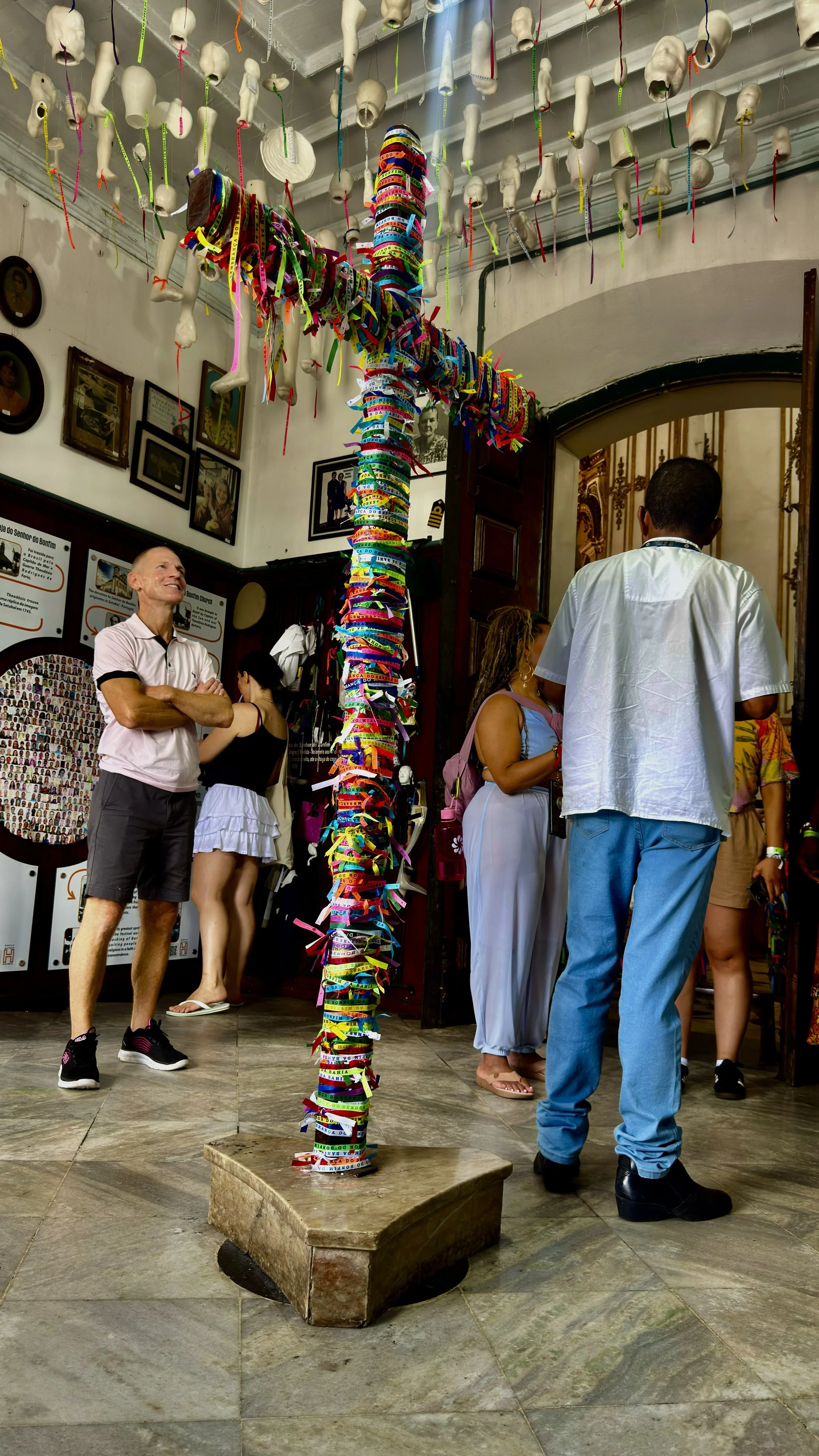 A group of people standing inside a museum, looking at a display of hanging baby doll heads and a tall pole wrapped with colorful ribbons.