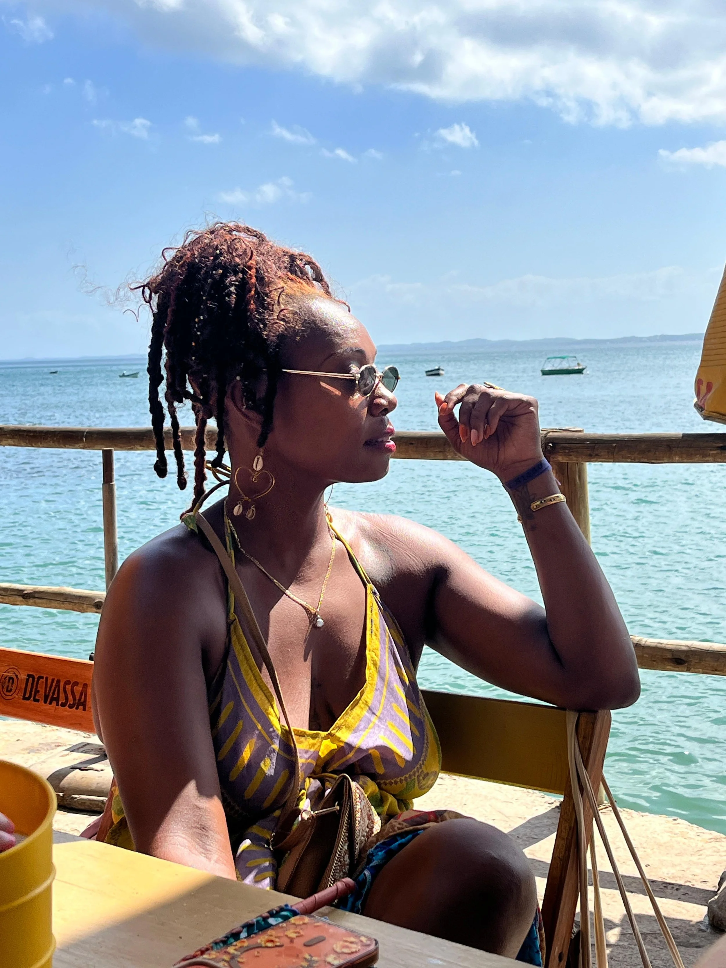 A woman with dreadlocks wearing sunglasses and jewelry sitting outdoors near the water, with boats and a blue sky in the background.