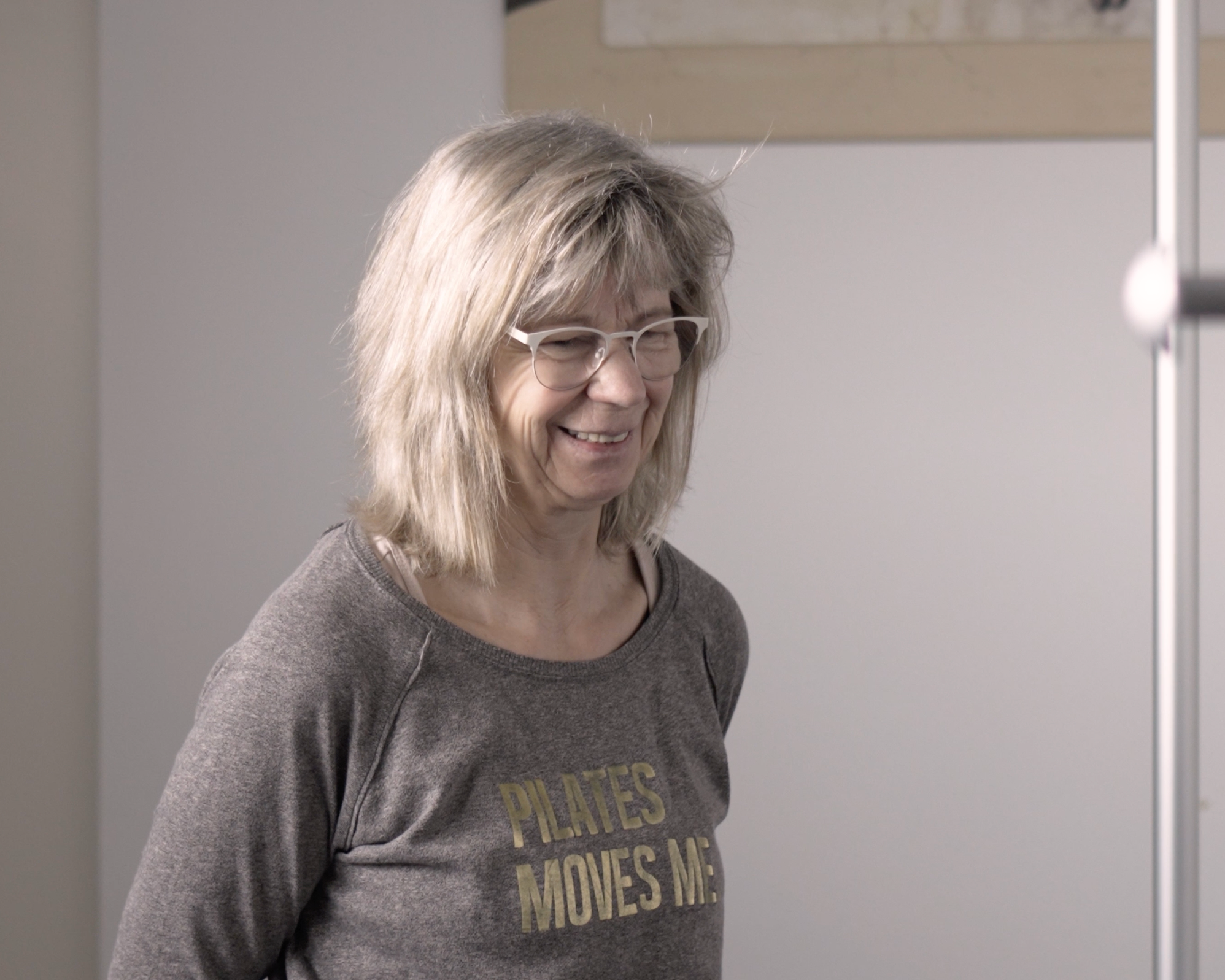 Une femme à cheveux gris clair, portant des lunettes rondes et un sweat gris avec des mots en français, sourit dans un environnement intérieur.