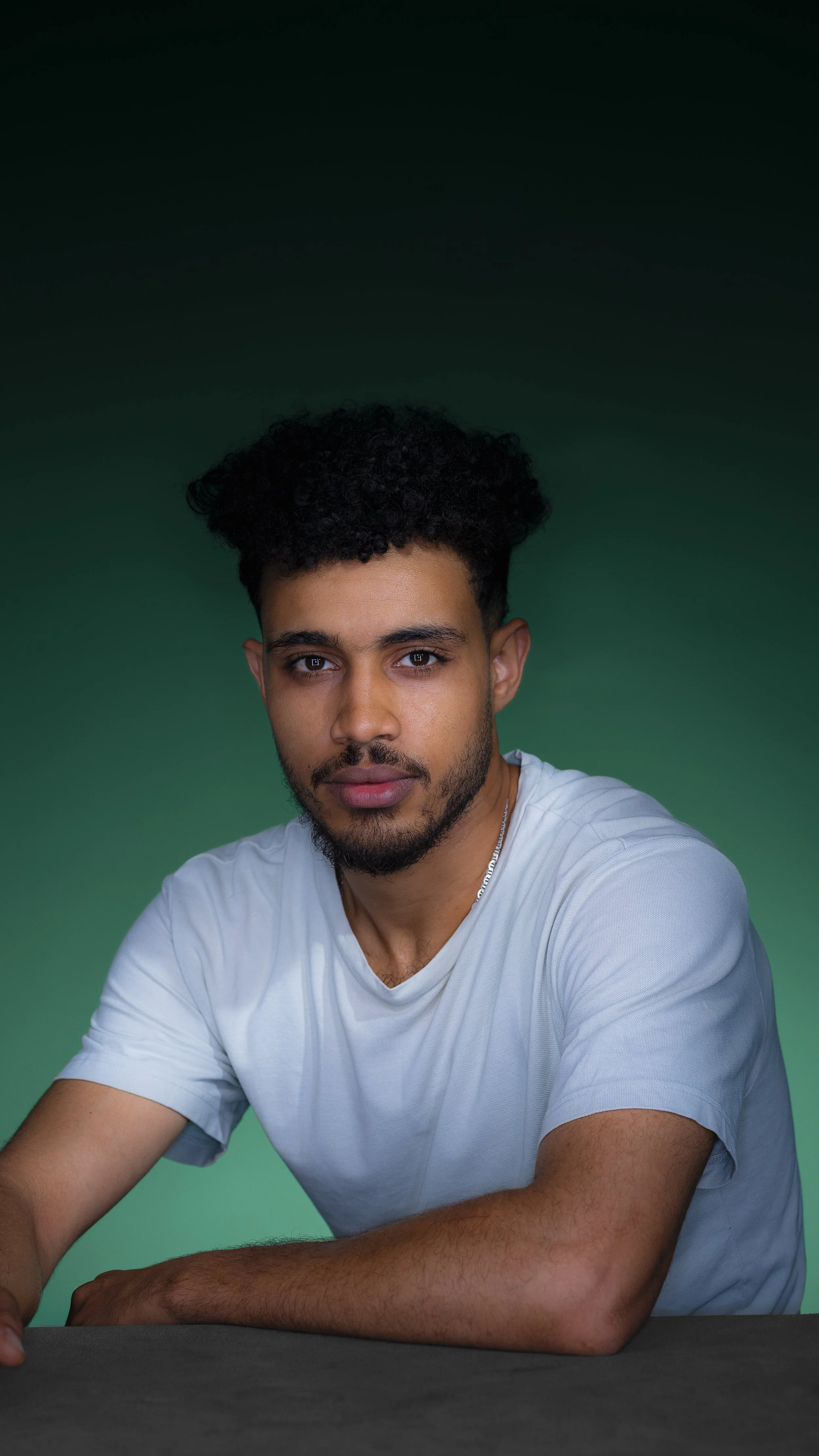 Young man with dark, curly hair and a beard, wearing a light gray t-shirt, sitting at a table against a green and black gradient background.