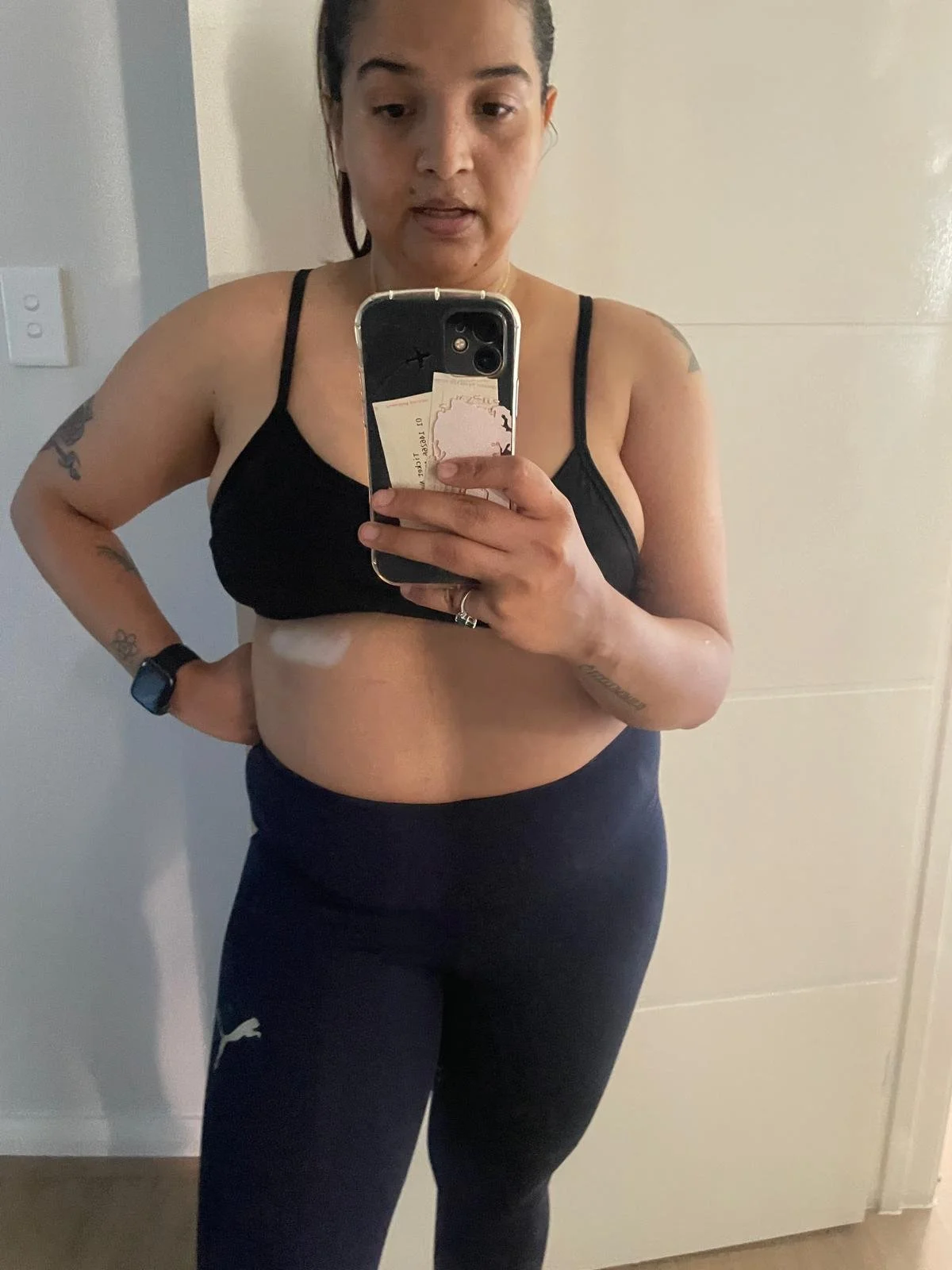 Woman taking a mirror selfie wearing a black sports bra and black leggings, with tattoos on her left arm and wristwatch on her left wrist.