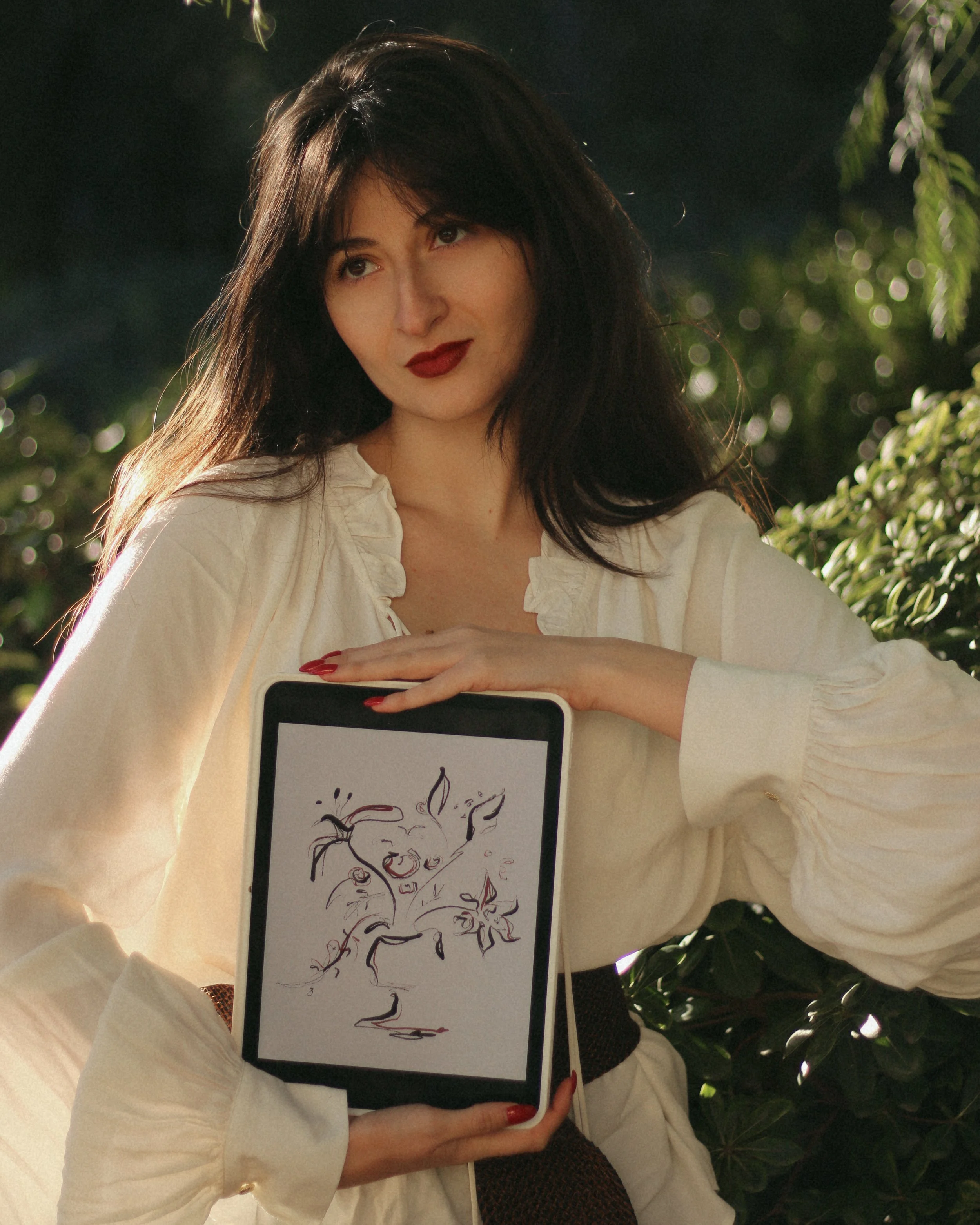 A woman with dark hair and red lipstick holding a tablet with a sketch of a face on it, outside near green foliage.