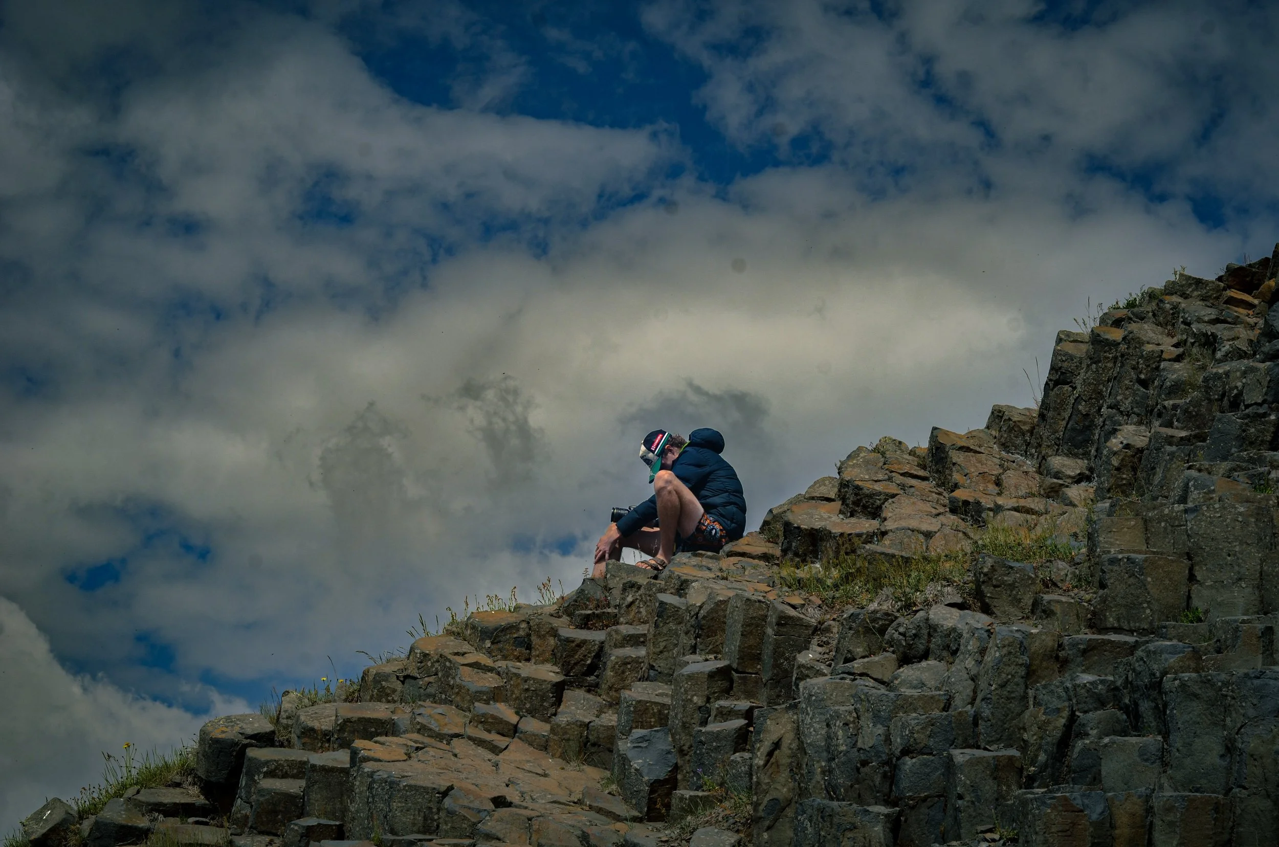 A person wearing a dark jacket, shorts, and a helmet crouches on a rocky mountain slope, with clouds and blue sky in the background.