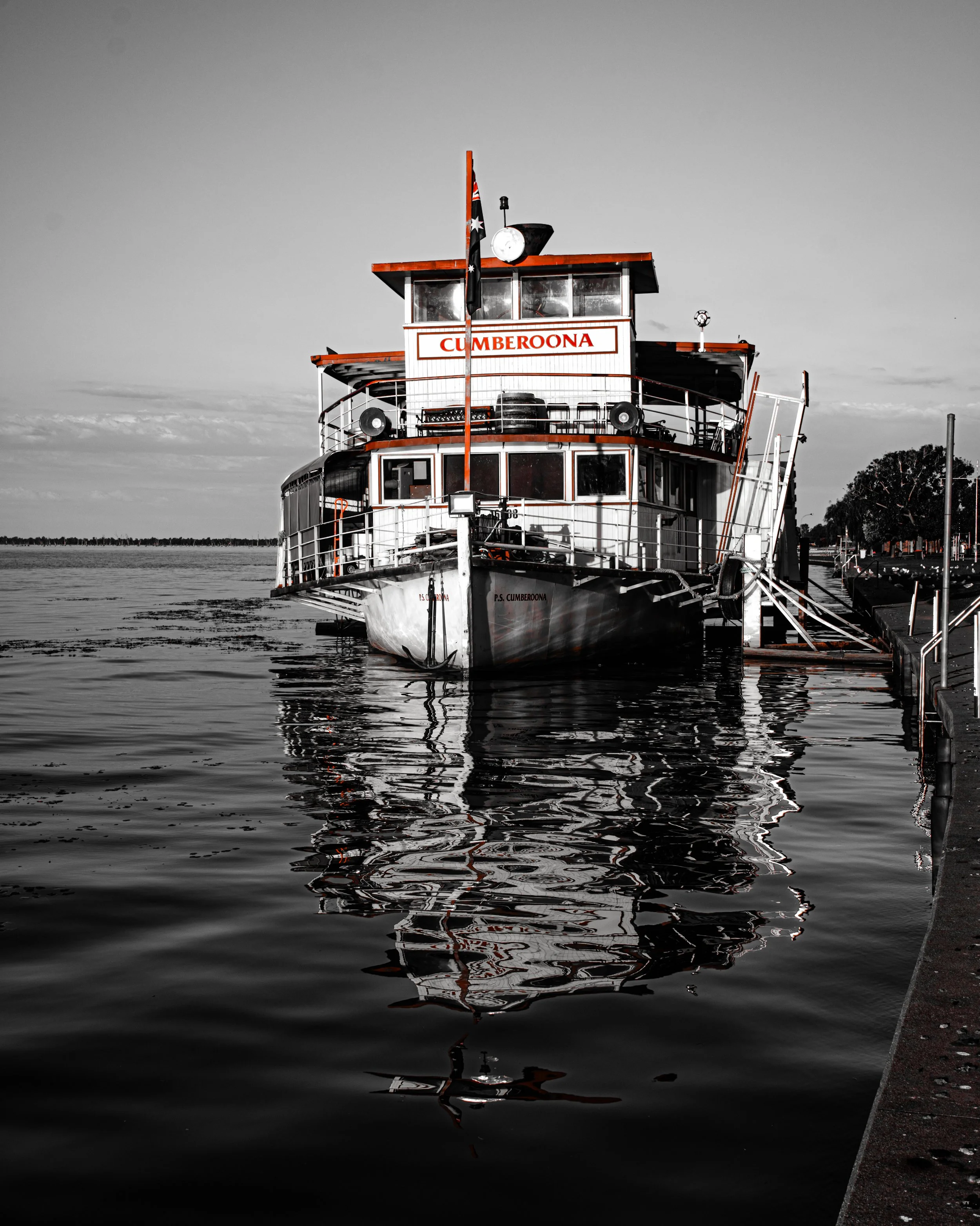 A black and white photograph of a boat docked at a pier, with the boat's name 'CUMBEROONA' visible on the front. The boat is reflected on the calm water, and there is a flag flying at the top of the mast. Trees are seen in the background under a partly cloudy sky.
