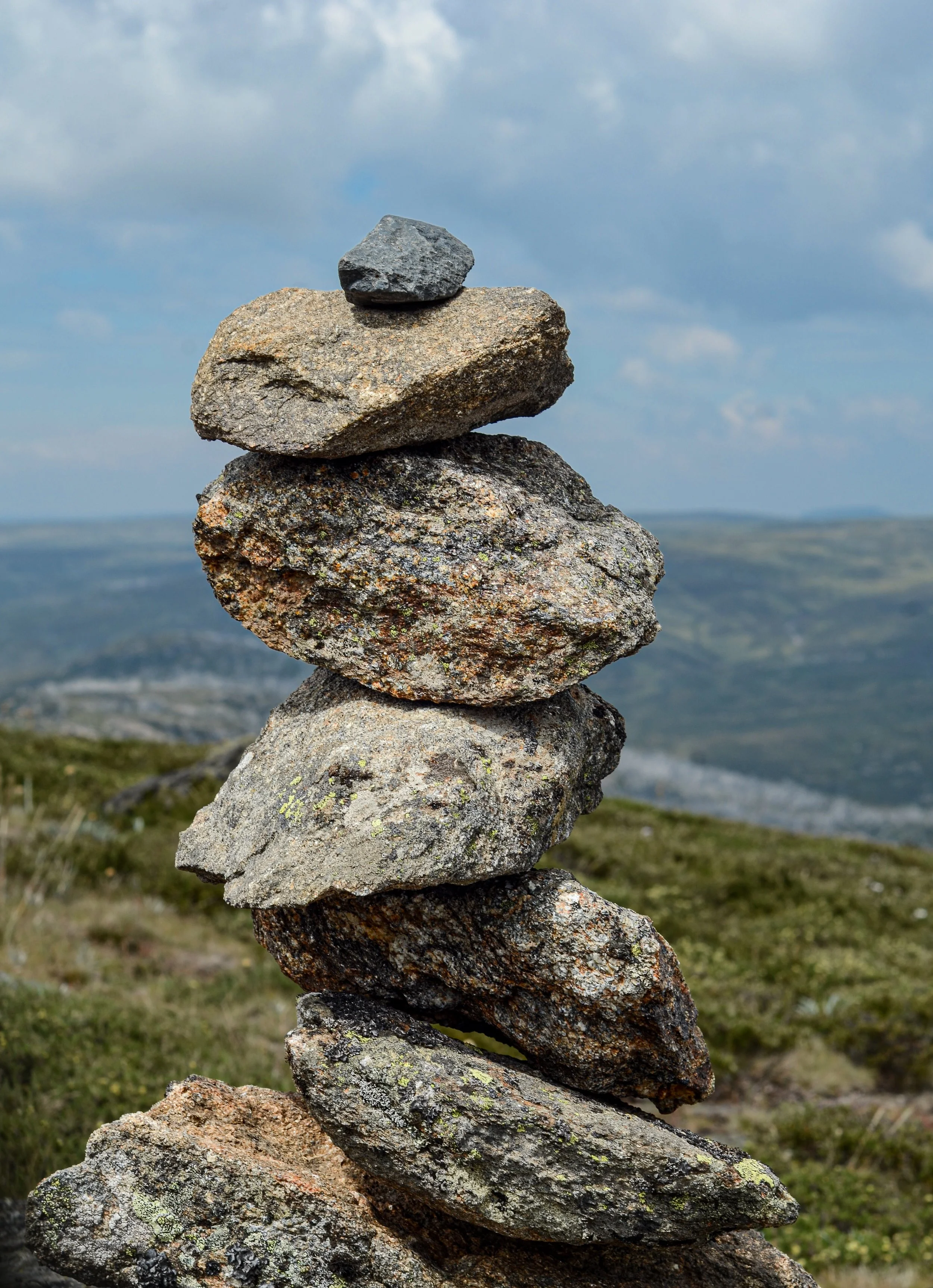 A vertical stack of rocks Balanced on top of each other with a cloudy sky and hilly landscape in the background.