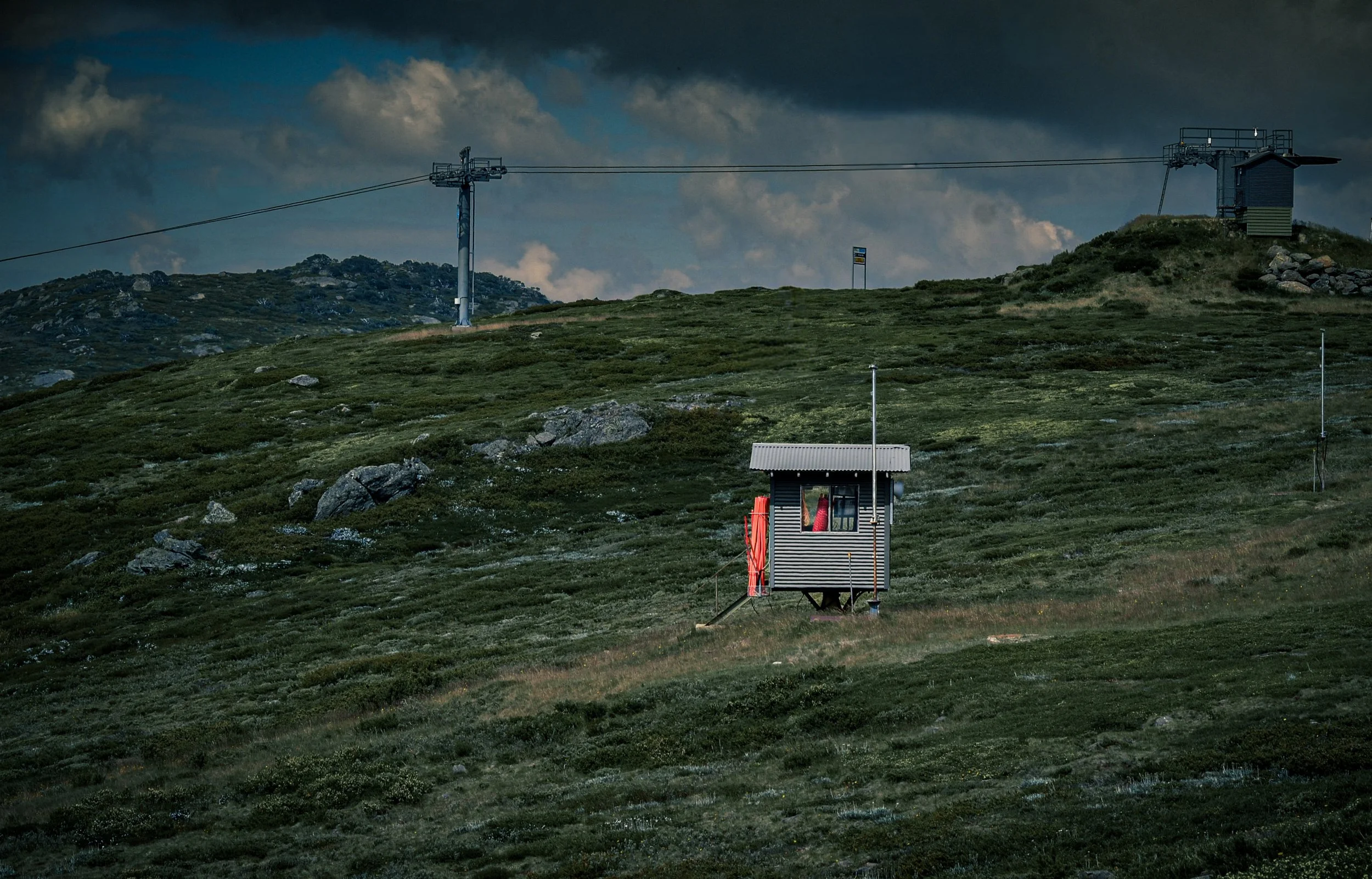 A mountain landscape with a ski lift, a small building, and a ski patrol hut under a cloudy sky.