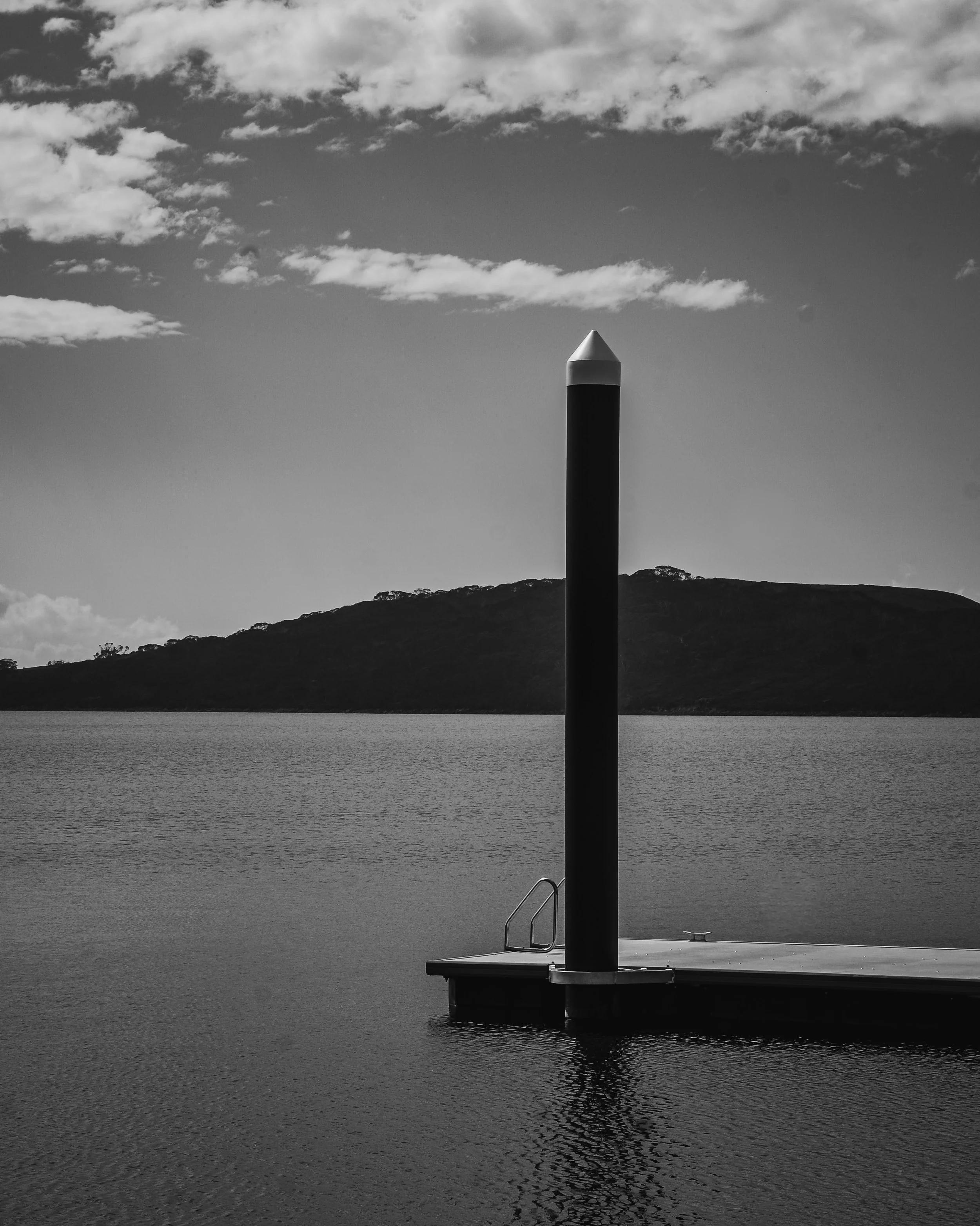 Black and white photo of a dock with a tall, narrow pole. The dock extends over calm water, with a mountainous horizon in the background and clouds in the sky.